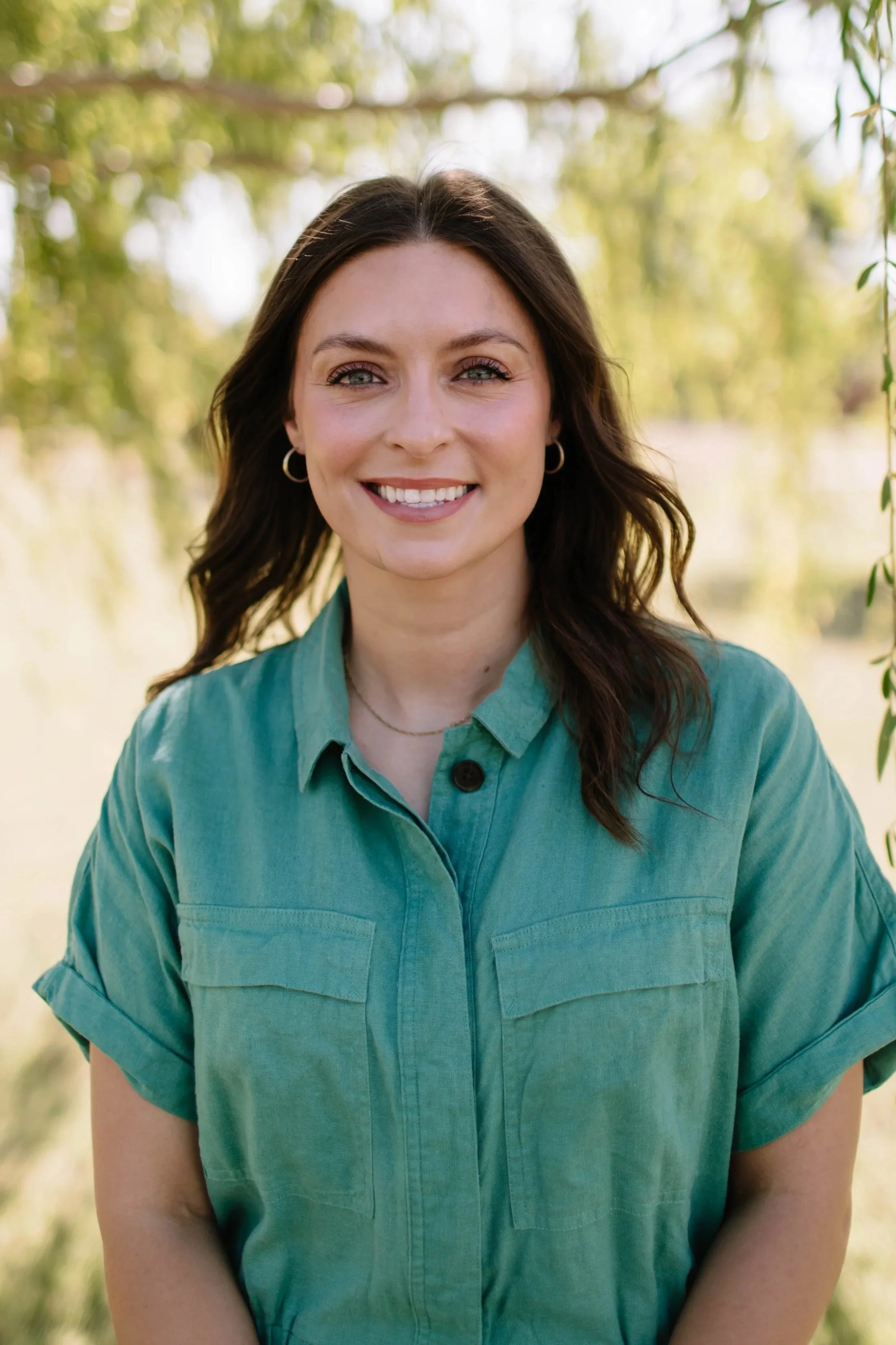 A woman with shoulder-length brown hair and blue eyes smiling outdoors, wearing a denim jacket and green earrings, with trees in the background.