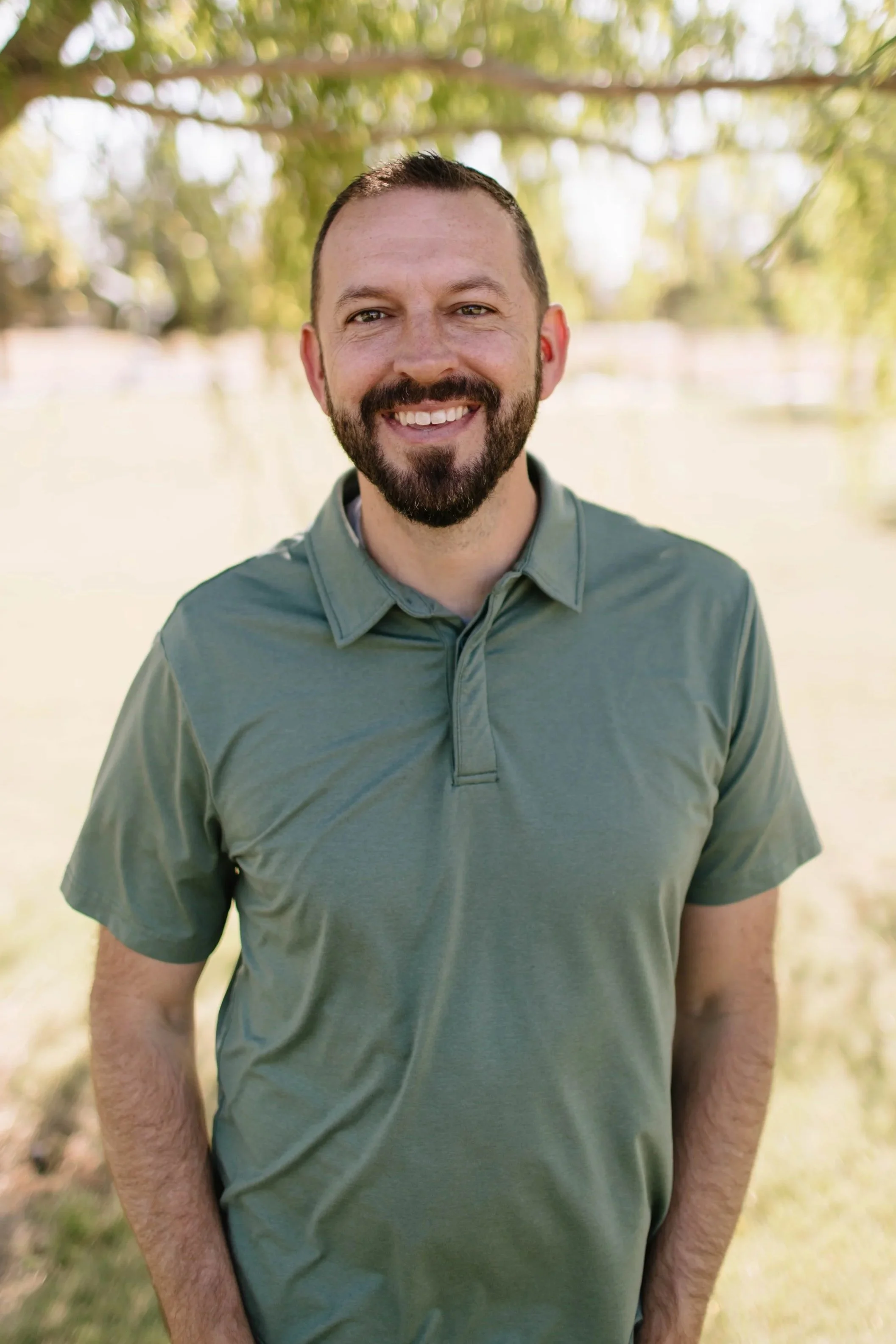 A man with a beard and short hair smiling outdoors, wearing a blue, white, and tan plaid shirt.