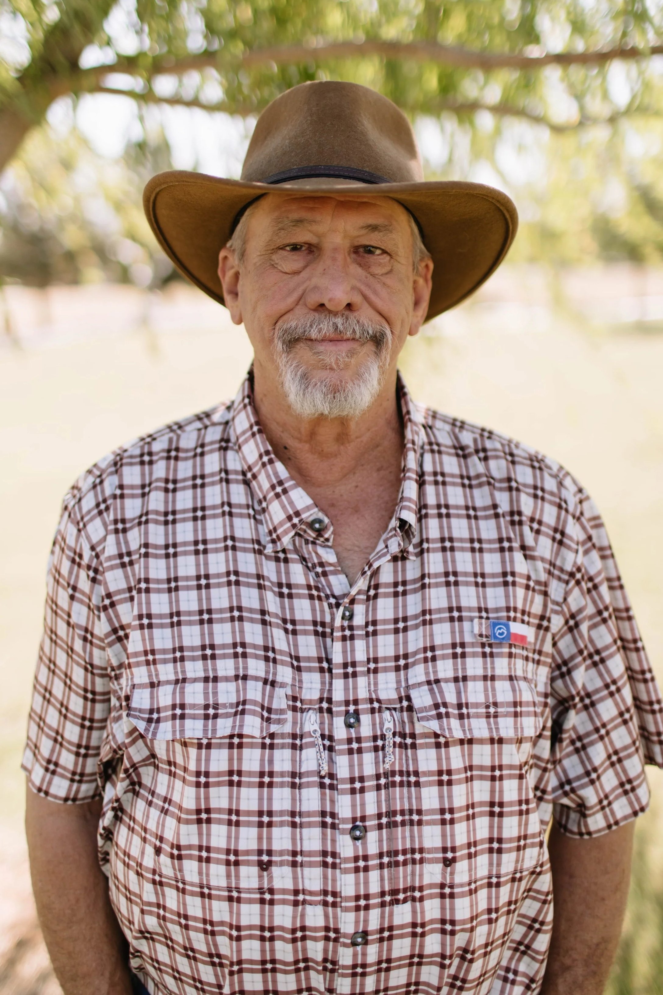 A man with a gray beard and mustache wearing a cowboy hat, a light-colored vest over a plaid shirt, and a neck gaiter, standing outdoors in front of a brick wall and greenery.