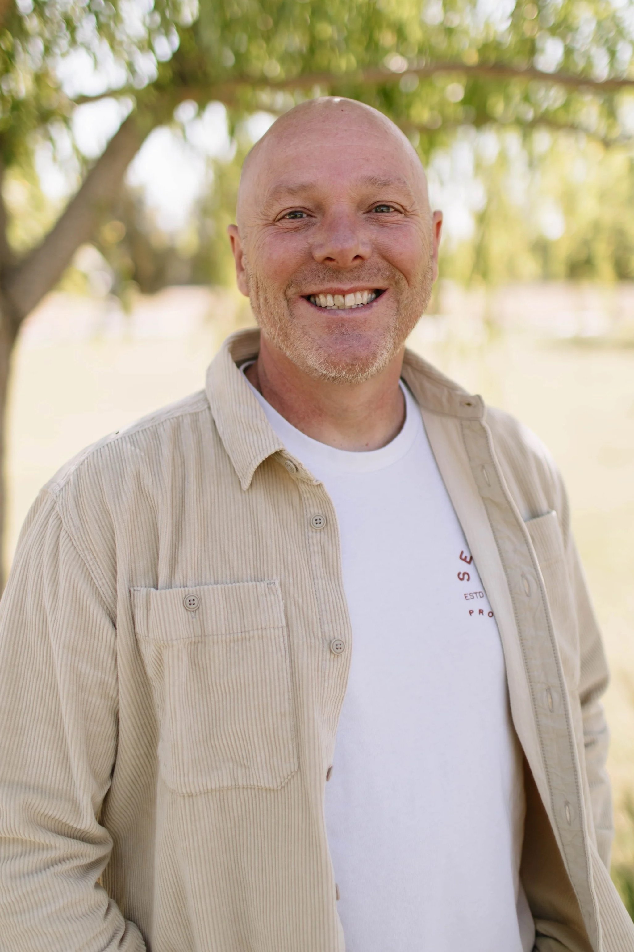 A smiling man with a bald head and light beard, wearing a black polo shirt with a small red logo, standing outdoors in front of a large tree with autumn foliage.
