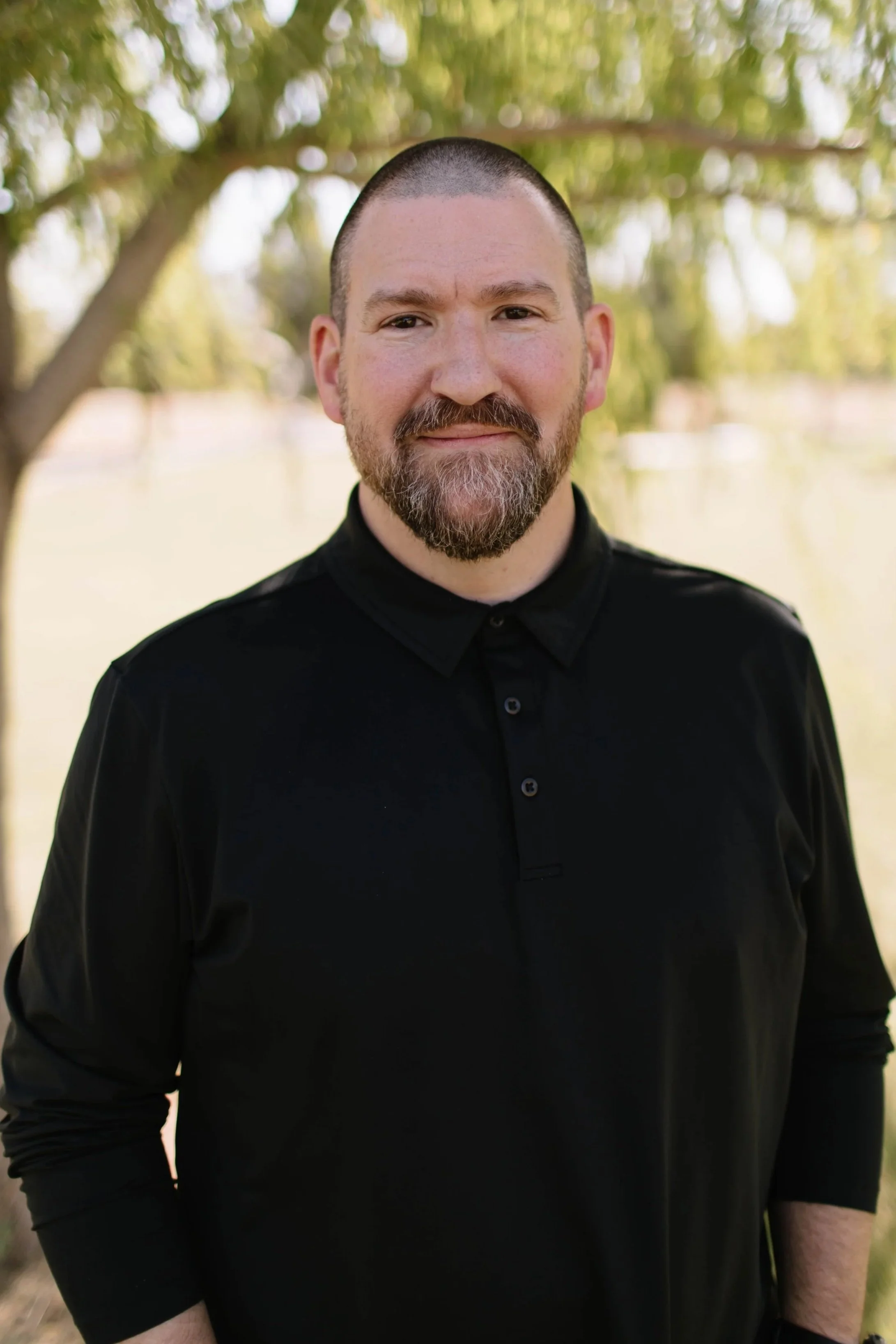 A smiling man with a beard and short hair standing outdoors in front of trees, wearing a dark gray cardigan and a blue striped button-up shirt.