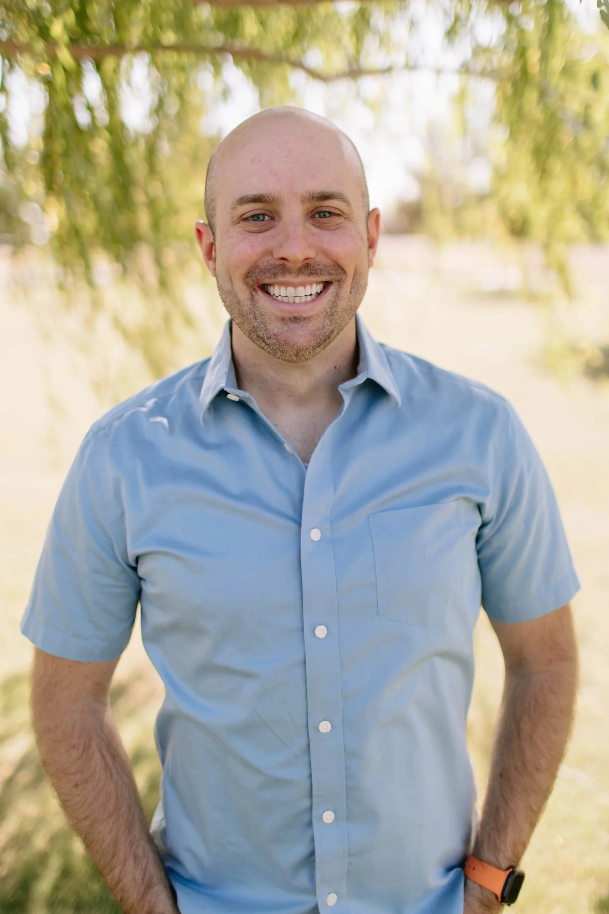 A smiling man with a beard and short hair, wearing a pink and white checkered shirt, outdoors with trees and sunlight in the background.