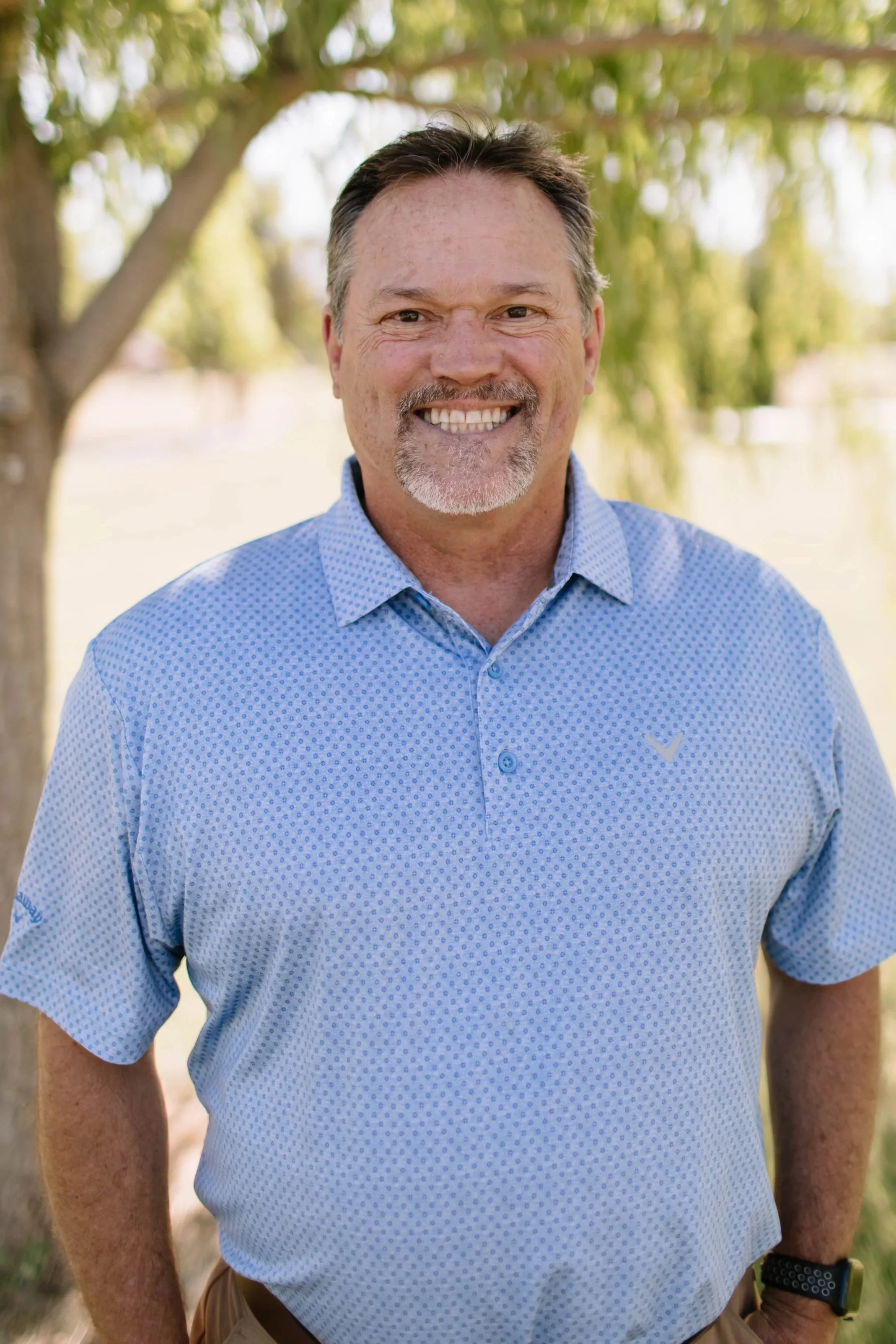 A middle-aged man with short brown hair, a goatee, and a tan complexion smiling outdoors. He is wearing a khaki button-up shirt with chest pockets, standing in front of trees and greenery.
