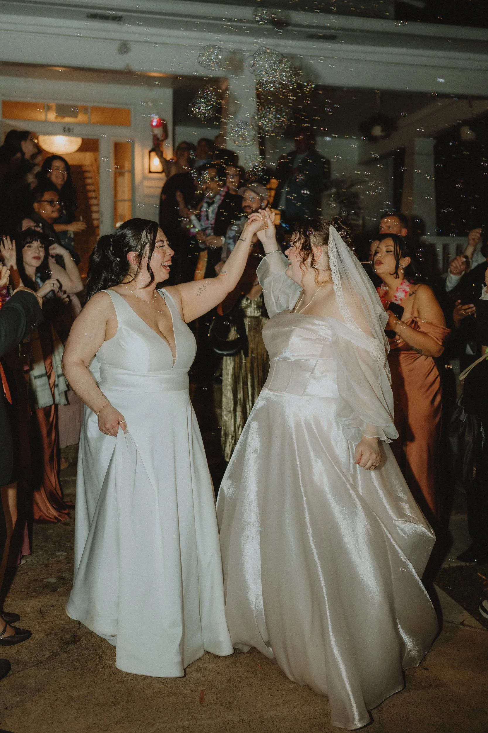 Two brides walking out of their wedding reception holding hands while surrounded by bubbles and guests