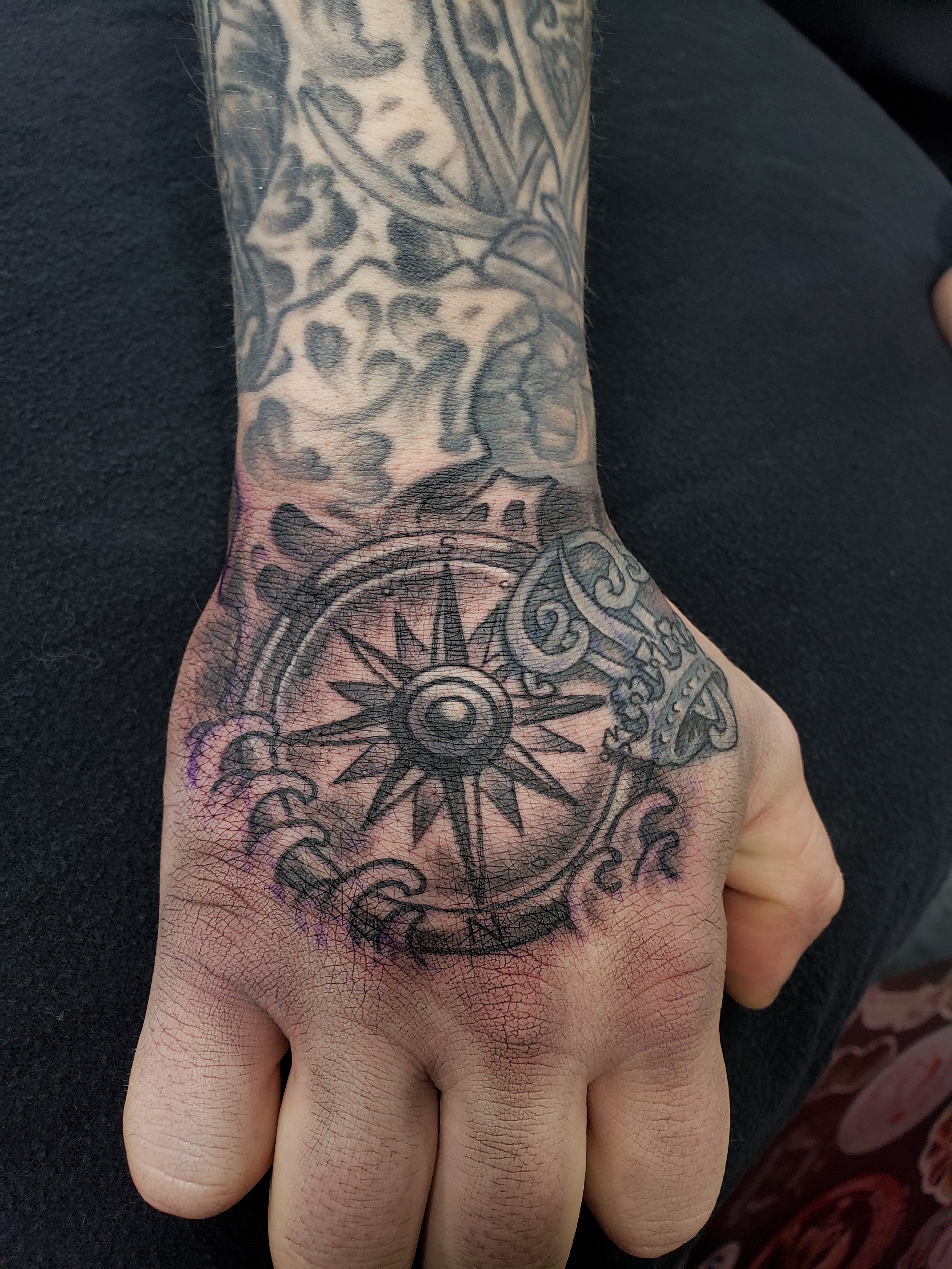 Tattoo on a person's hand and wrist featuring a large compass with cardinal directions and intricate designs.