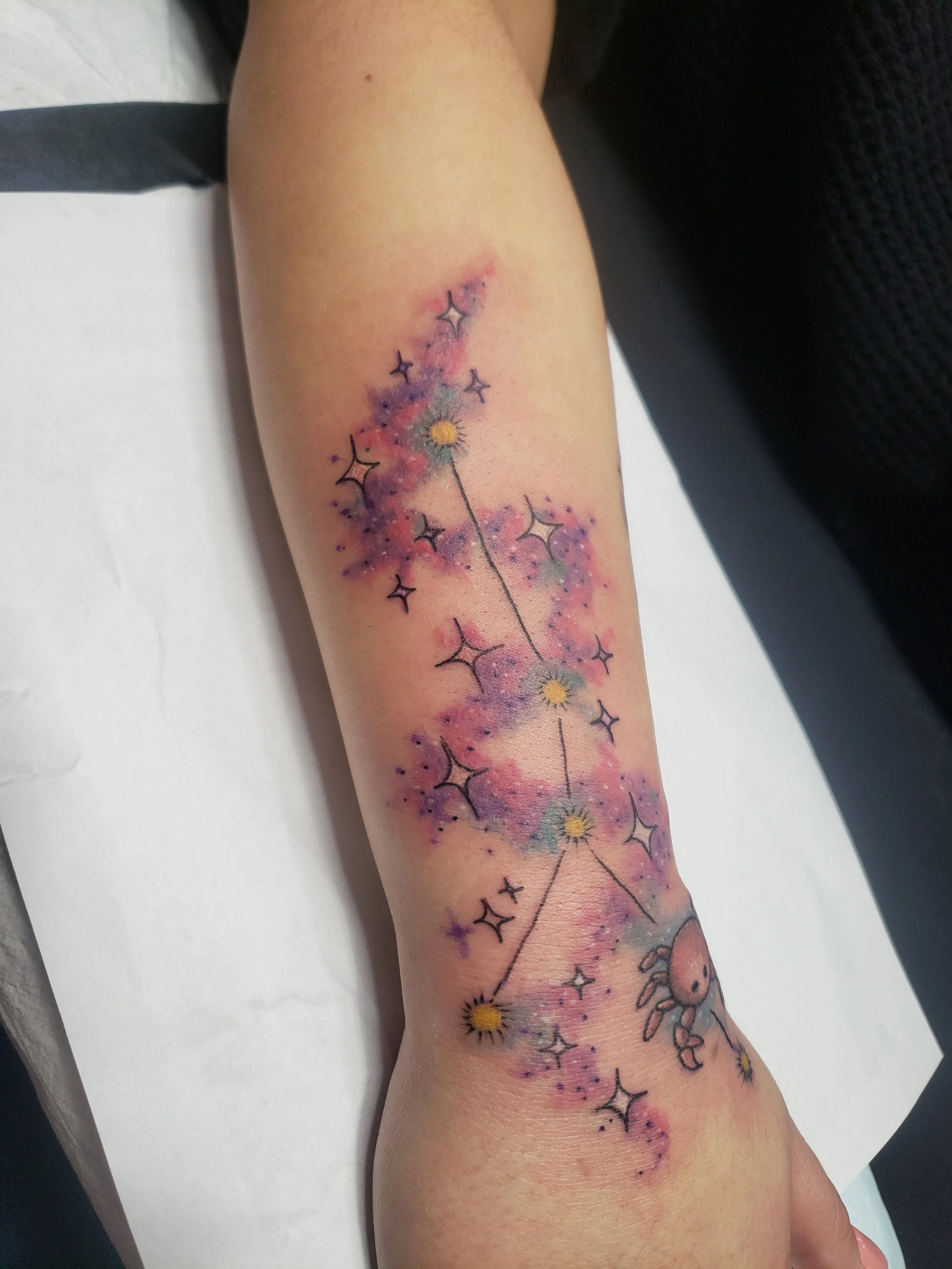 Colorful tattoo of a constellation with stars and a moon on a person's forearm, surrounded by watercolor-style pinks, purples, and yellow accents.