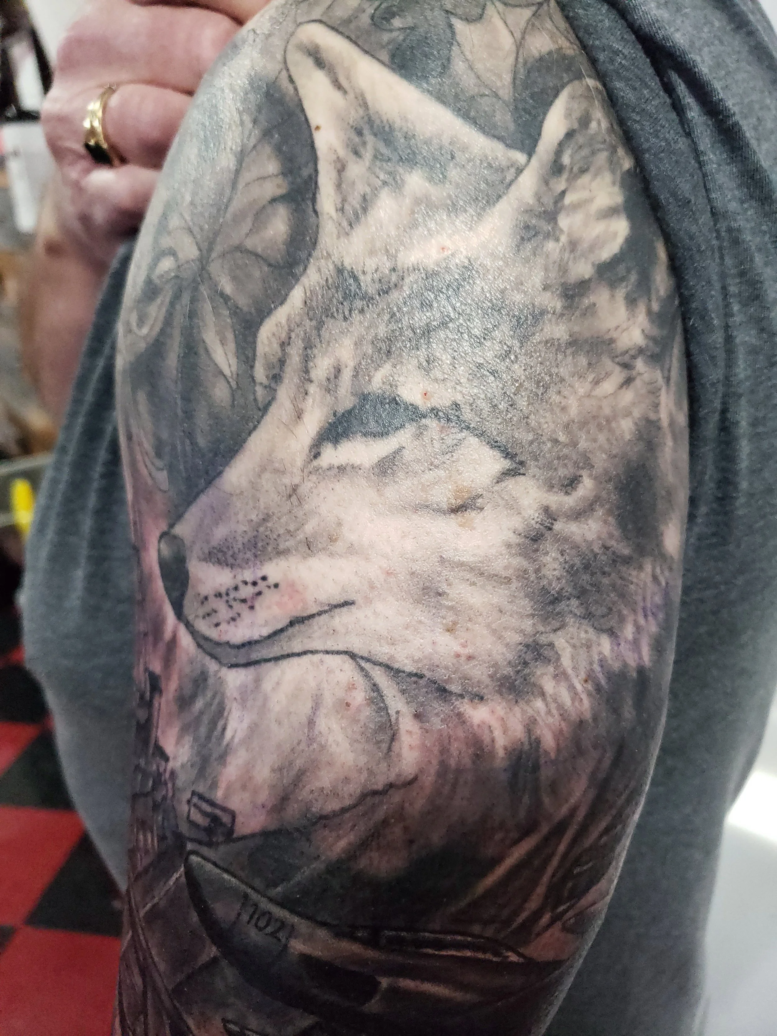 Close-up of a person's upper arm with a large, detailed black and gray tattoo of a dog's face, including the dog's nose, eyes, and mouth, surrounded by leaves and natural elements.
