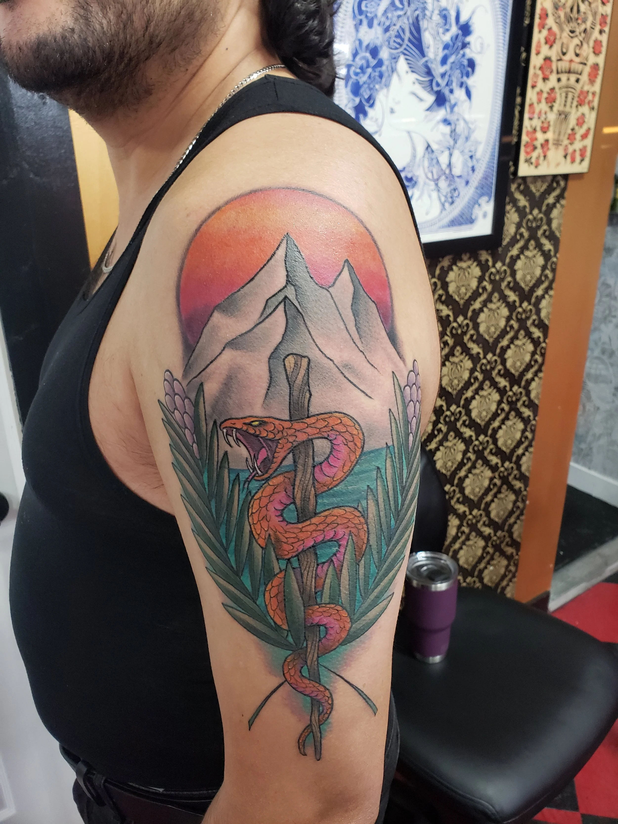 Tattoo of a snake intertwined with a mountain landscape, set against a pink and orange sunset background, on a person's upper arm.