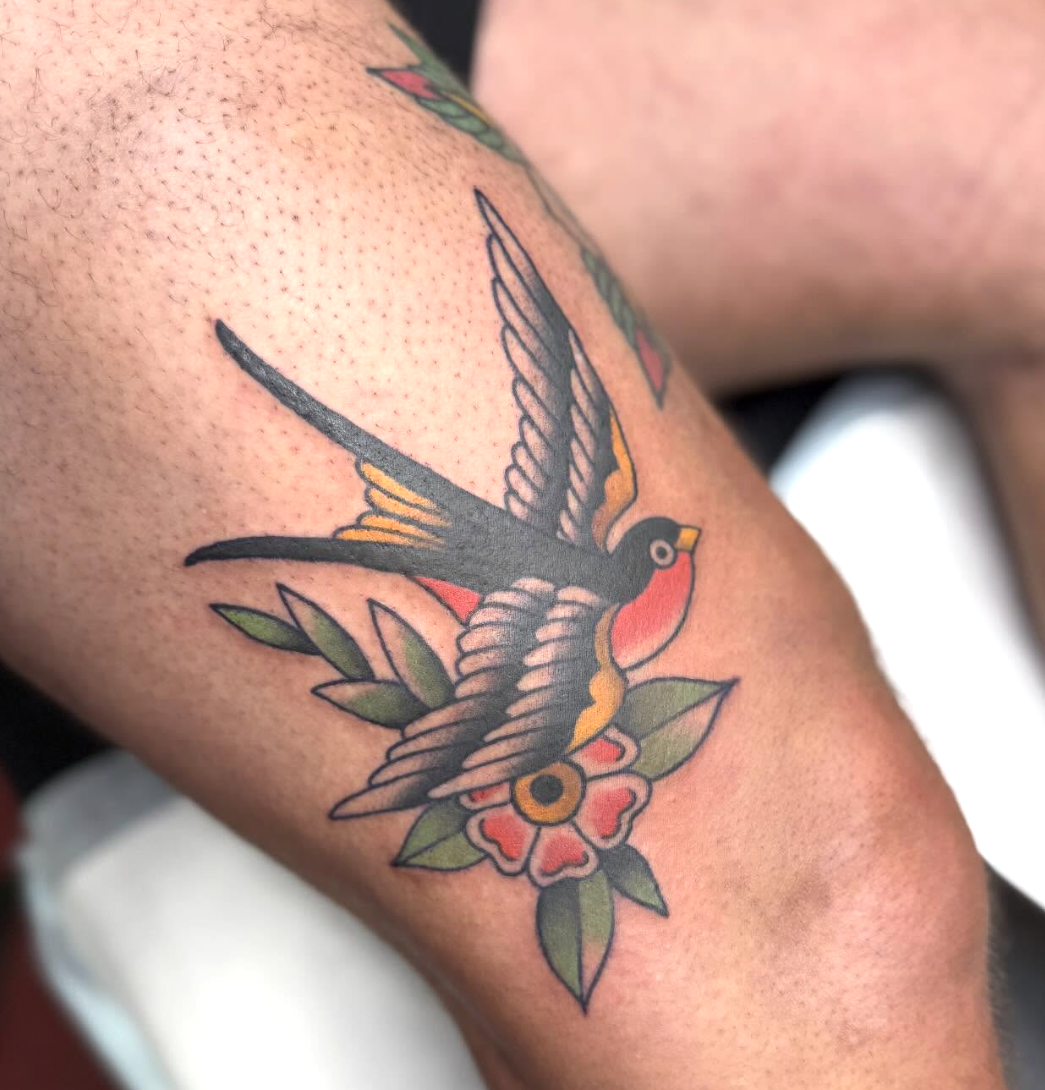 Traditional style tattoo of a swallow bird with a red and yellow chest, flying above a pink flower with green leaves.