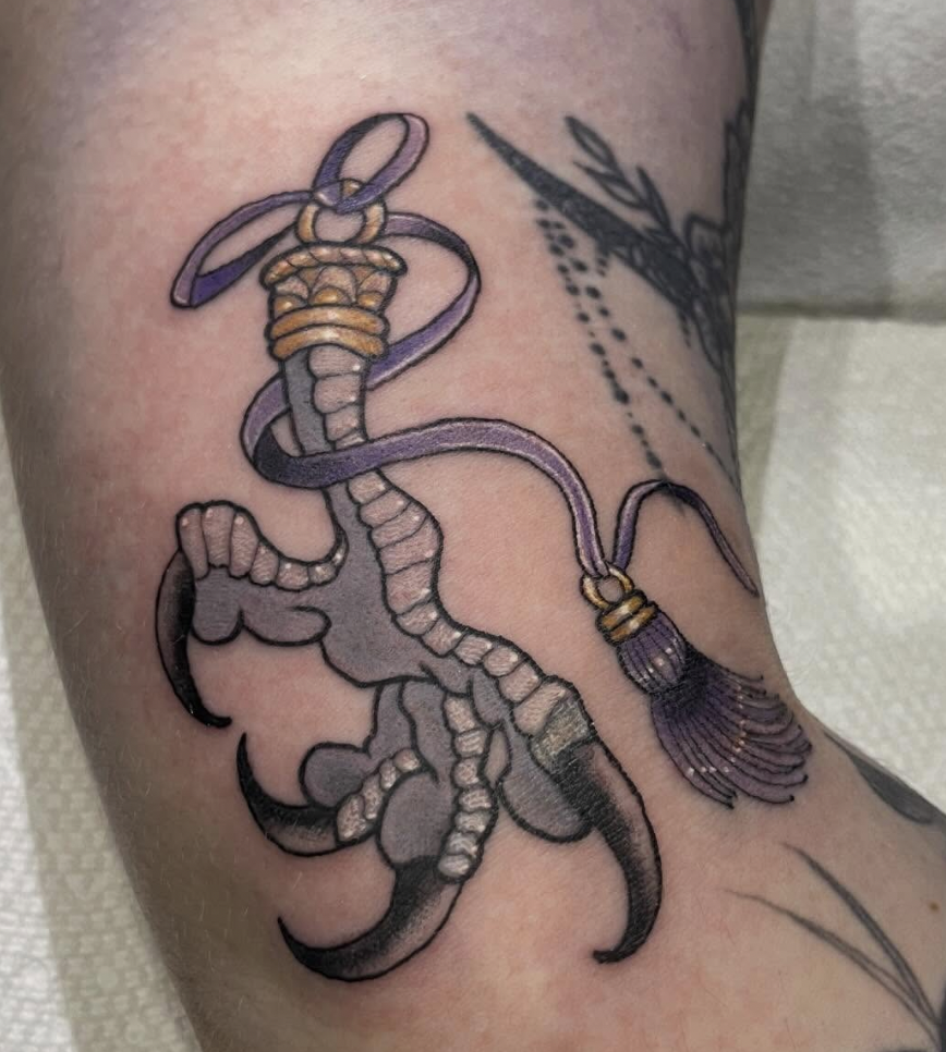 Tattoo of a scorpion with a purple electric plug and cord, with gold accents on the cord and a tassel on the plug.