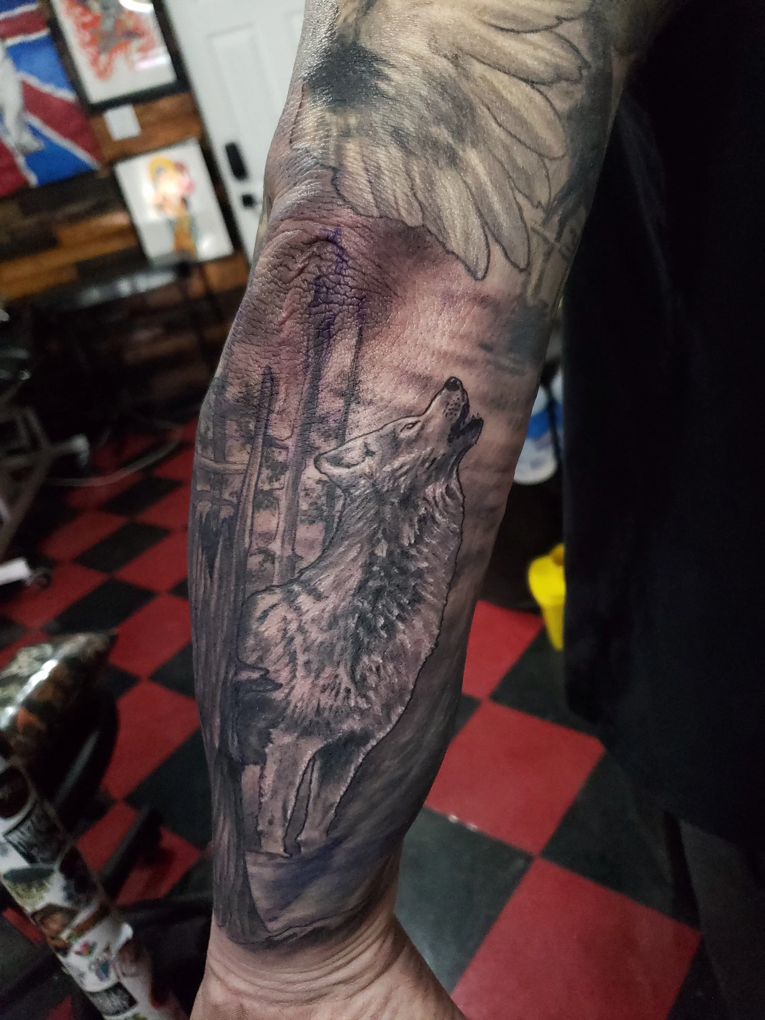 Tattooed arm featuring a wolf and trees with leaves and branches in black and gray ink.