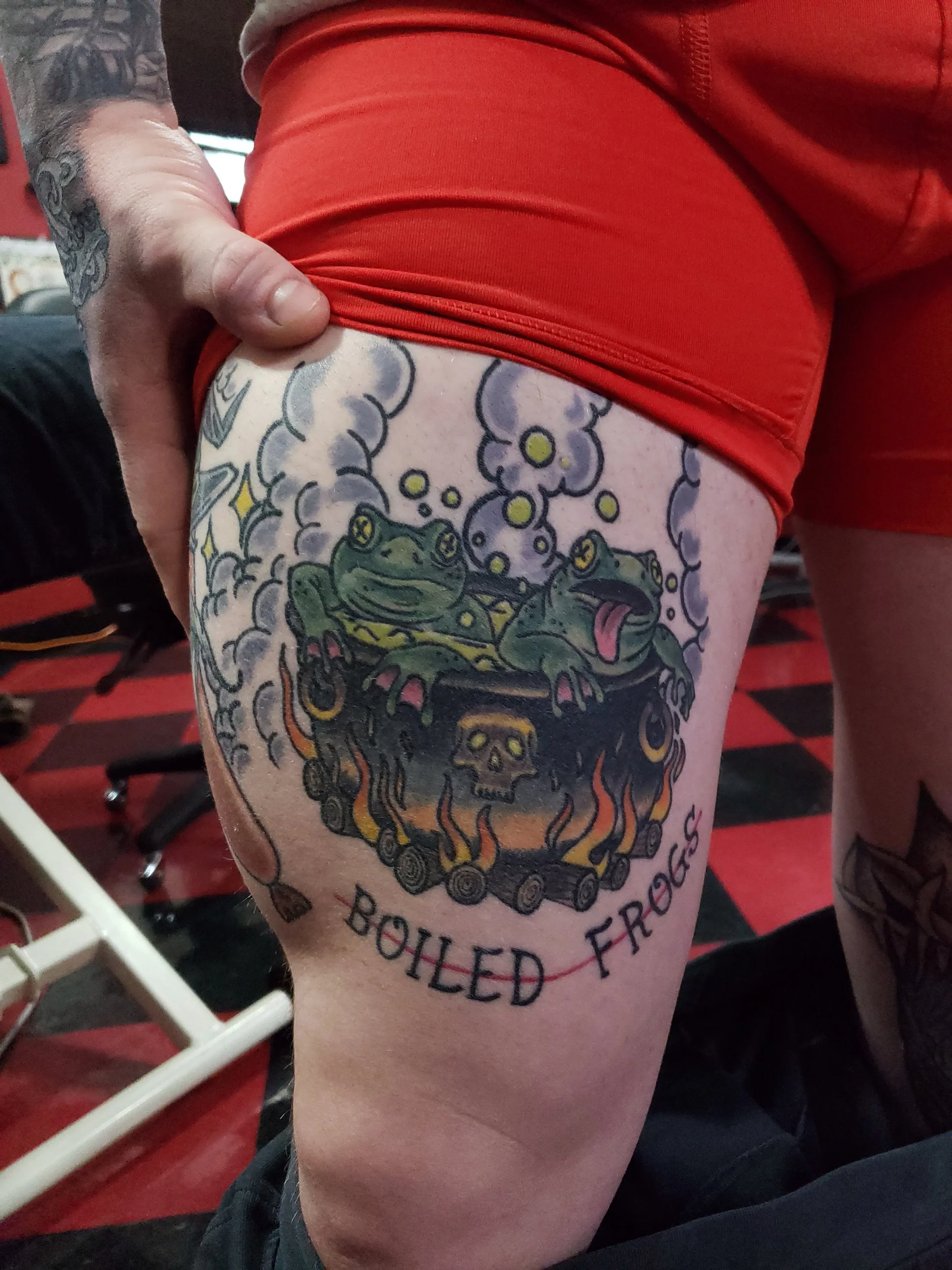 Tattoo of a green frog with a skull on its back, surrounded by purple smoke and flames, with the words "Boulder Frog" and "Frog Inside" on the thigh.