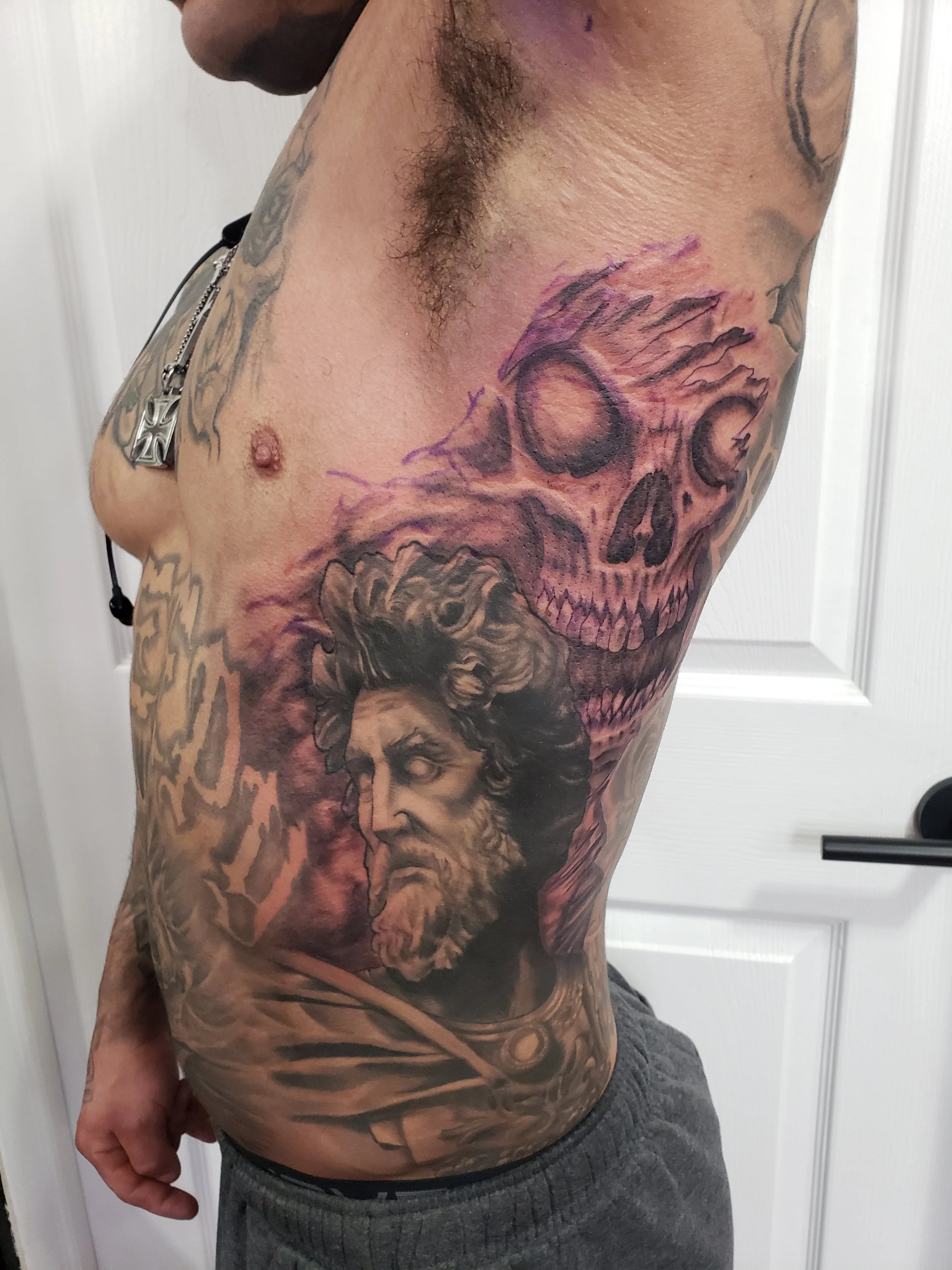 A person with tattoos on their upper arm and torso, standing sideways. The tattoos include a large skull with a woman's face on the side, and other designs.