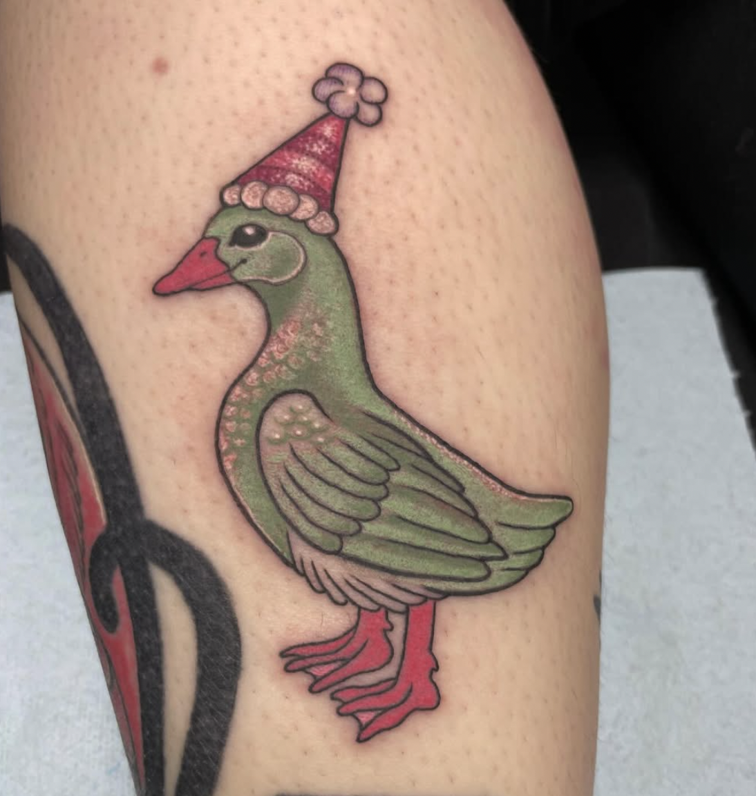 Tattoo of a cartoon duck wearing a red Santa hat with a pink pom-pom, on a person's arm.
