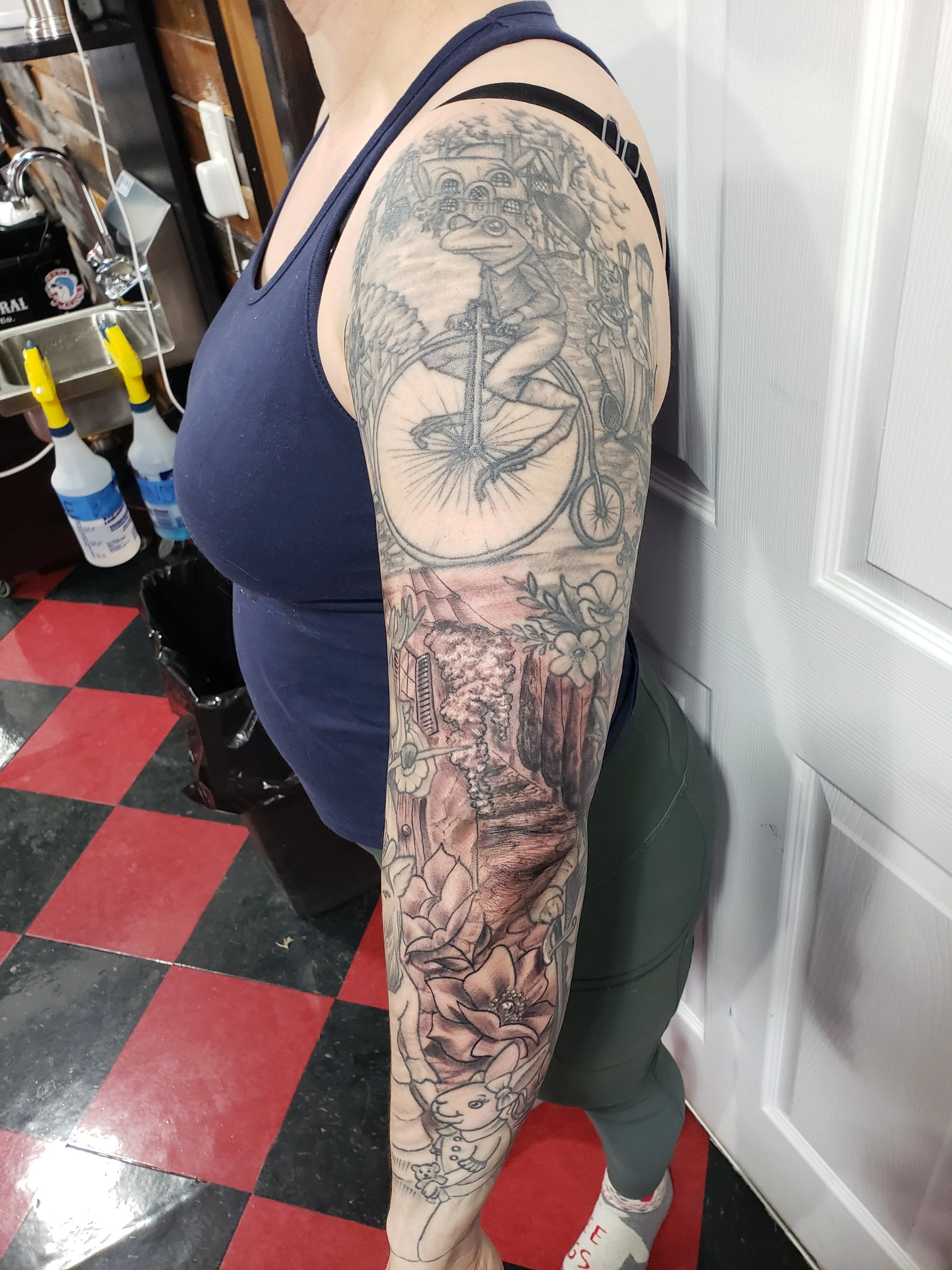 A person with a large detailed black and grey tattoo sleeve on their arm, featuring a frog cycling on a bike, a house, flowers, and characters, wearing a navy tank top, dark shorts, and socks, standing indoors on a red and black checkered floor.
