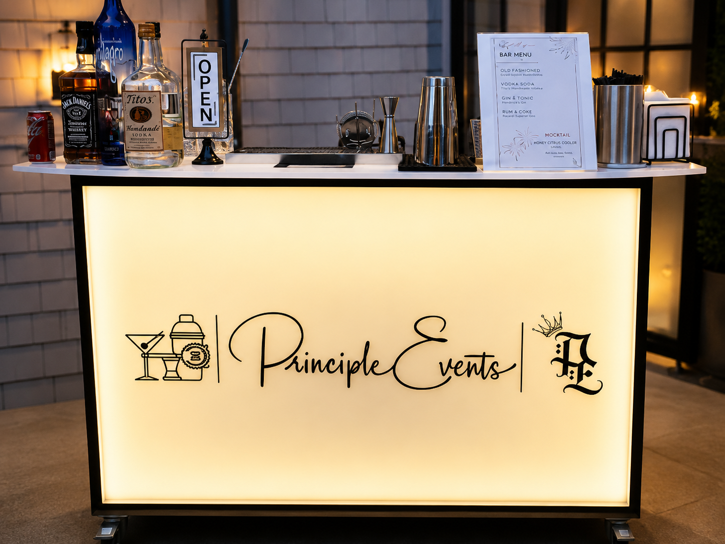 A illuminated bar counter with a sign reading "Principle Events". The counter has various liquor bottles, a cocktail sign, an open sign, a martini, and cocktail straws, with a printed menu and bar tools on top.