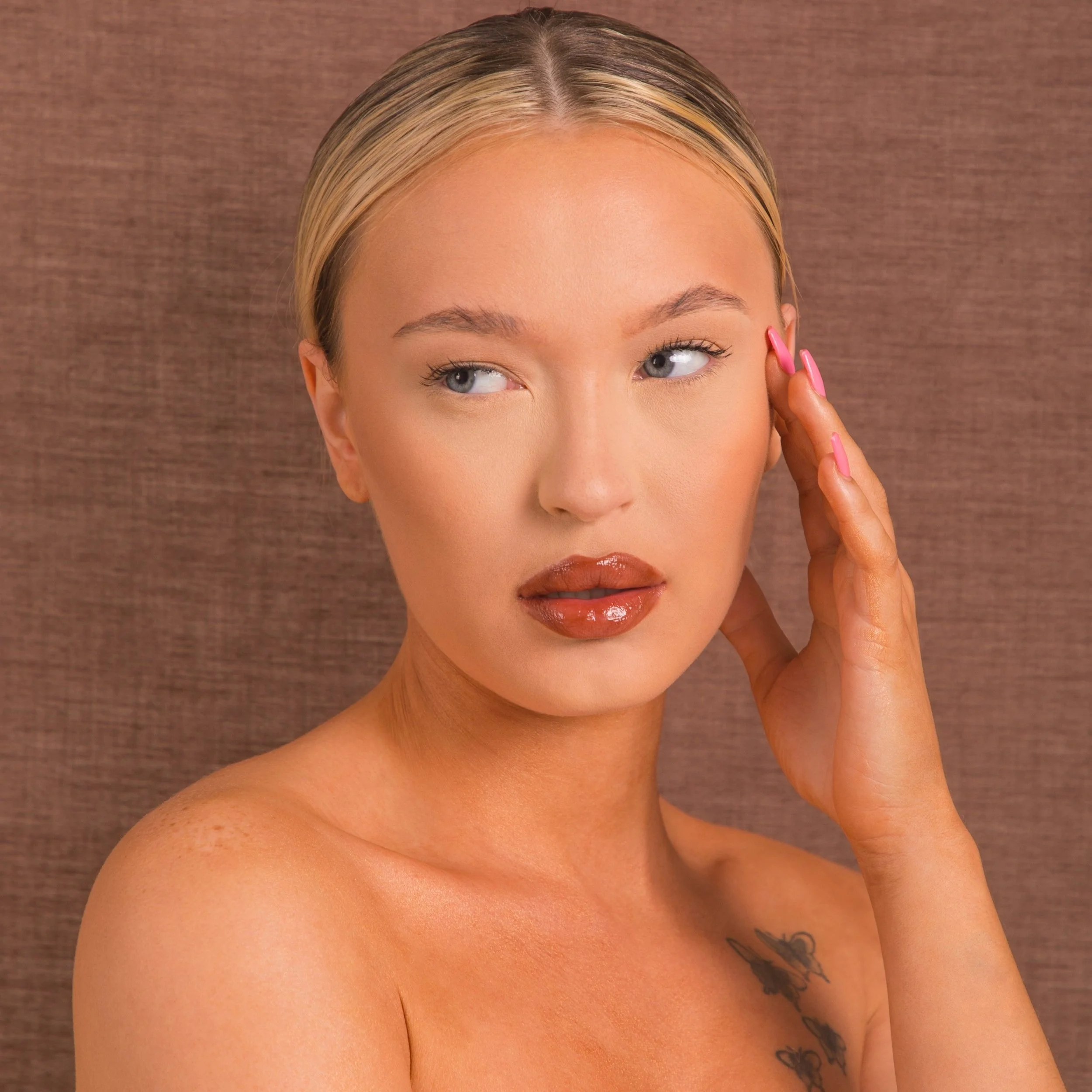 A young woman with blonde hair, blue eyes, and light skin touching her temple. She has glossy lips, painted nails, and a tattoo on her shoulder, set against a brown textured background.