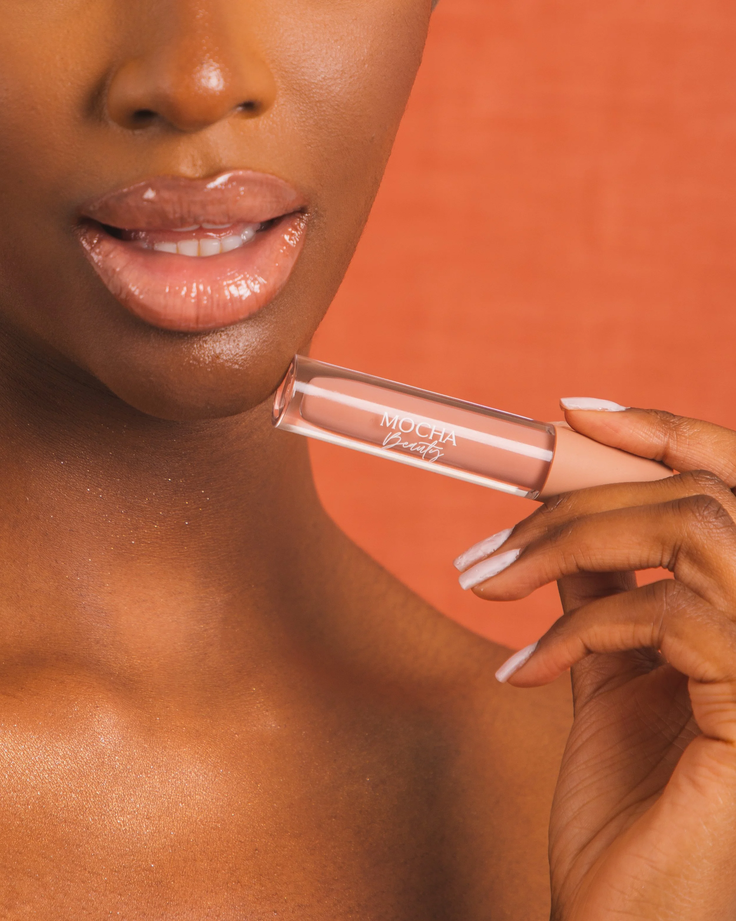 A woman with glossy lips holding a tube of Mocha beauty lip gloss near her mouth, with a peach-colored background.