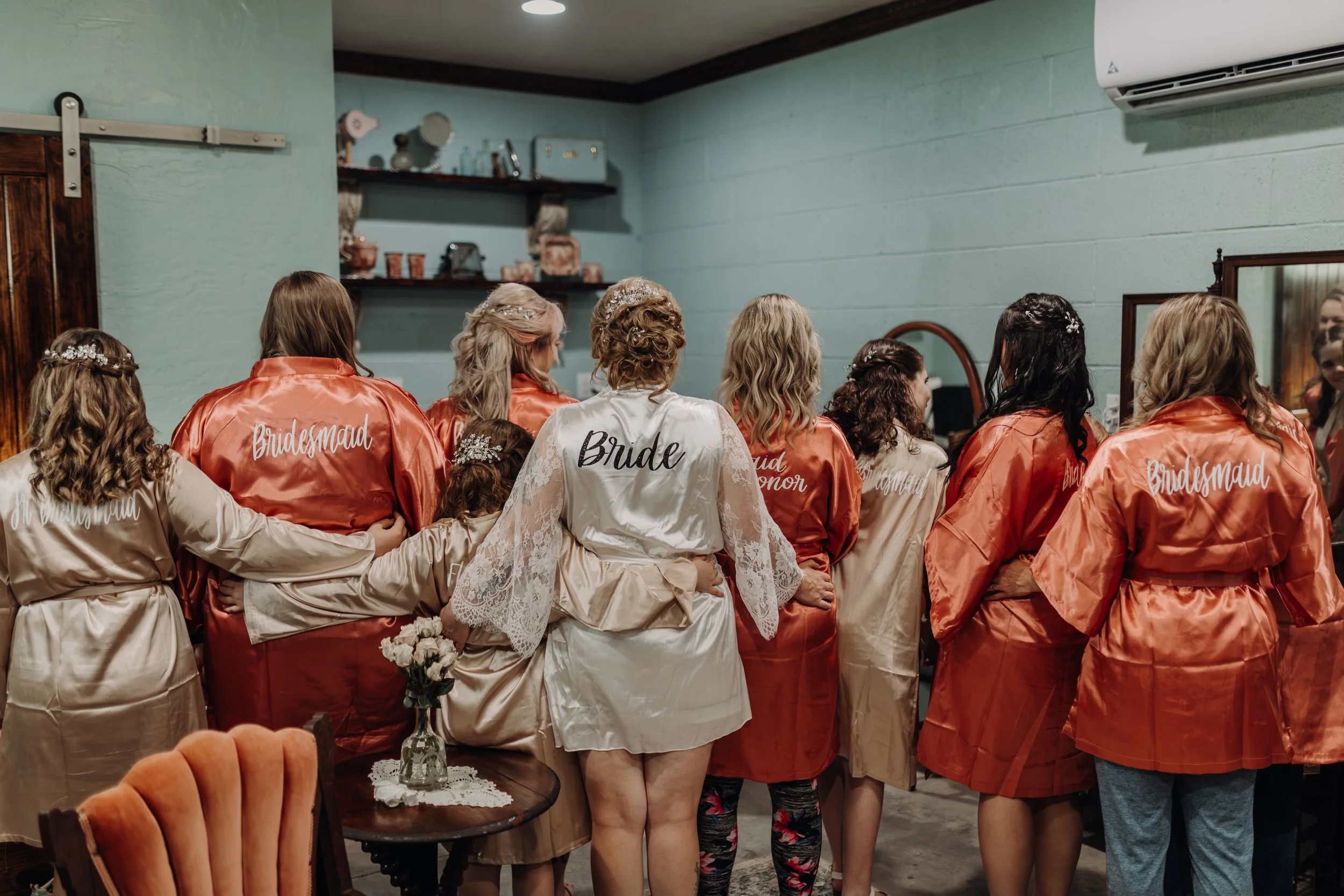 Bridal party wearing matching robes with 'Bride' and 'Bridesmaid' written on them, standing in a dressing room.