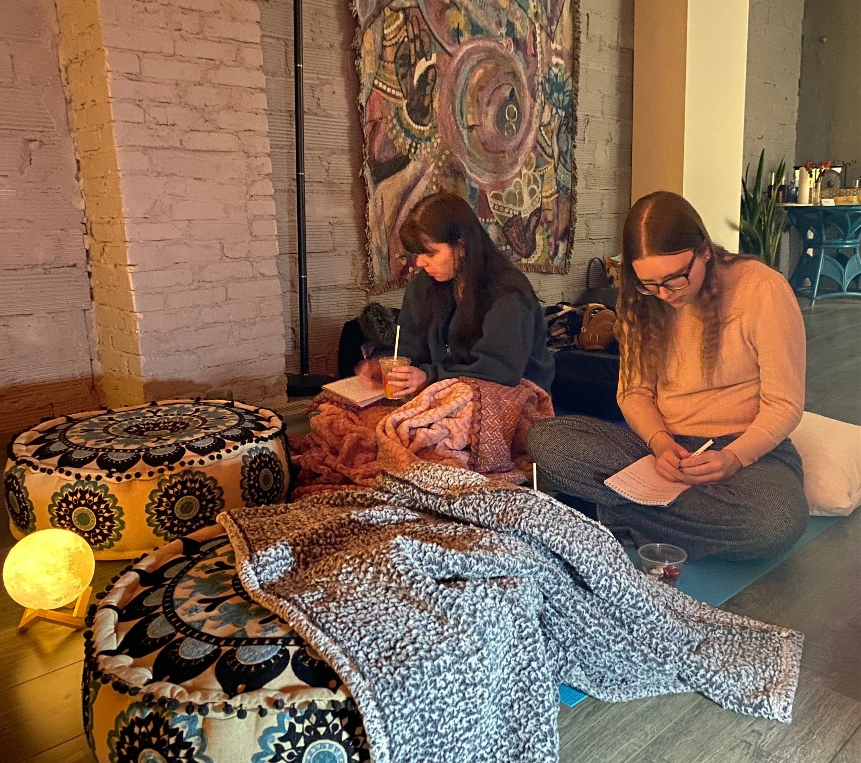 Two women sitting on cushions on the floor in a cozy room, engaged in writing or sketching, with blankets, a small lamp, and drinks around them.