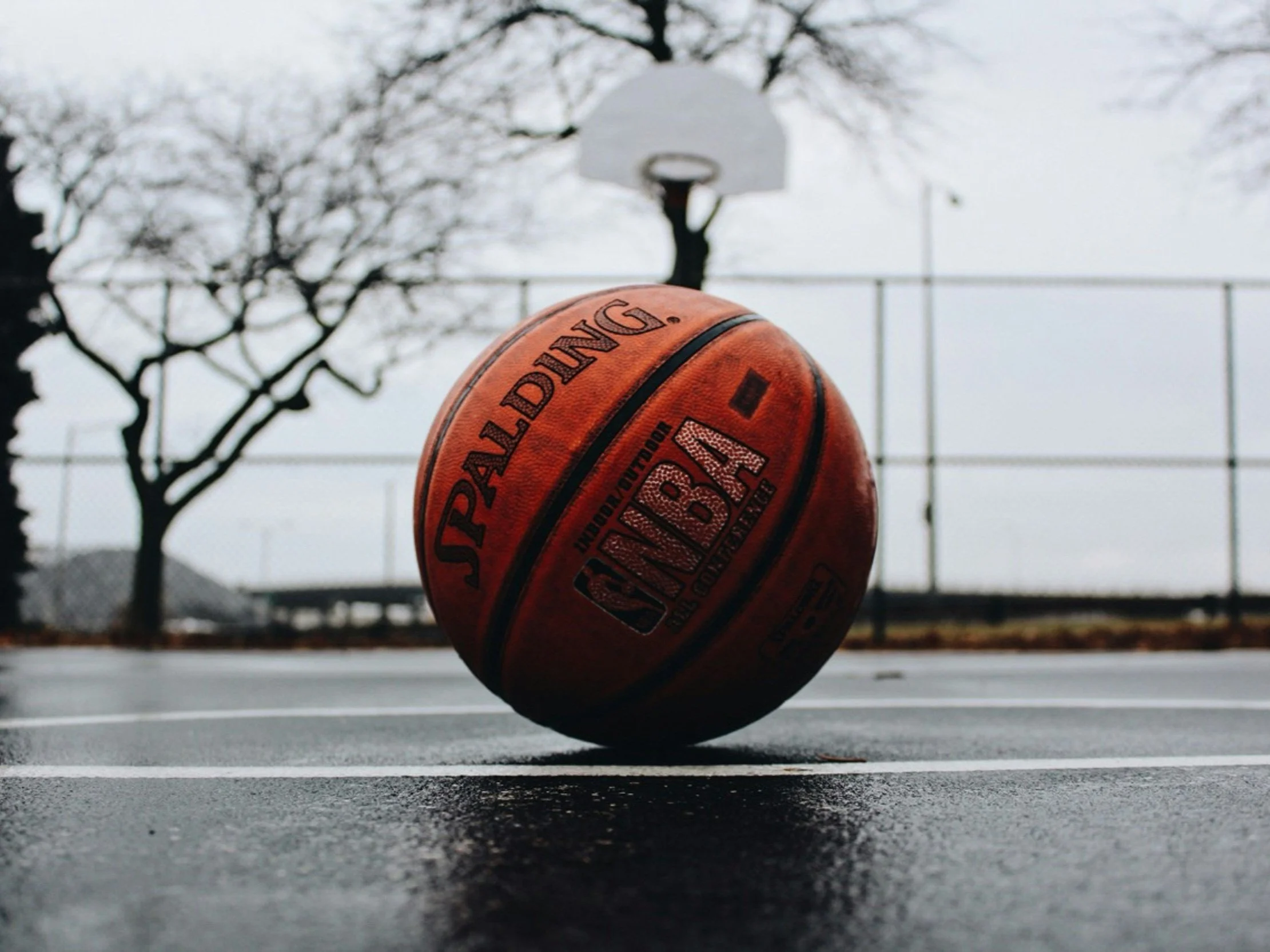 A basketball sits on top of the asphalt on a rainy day.