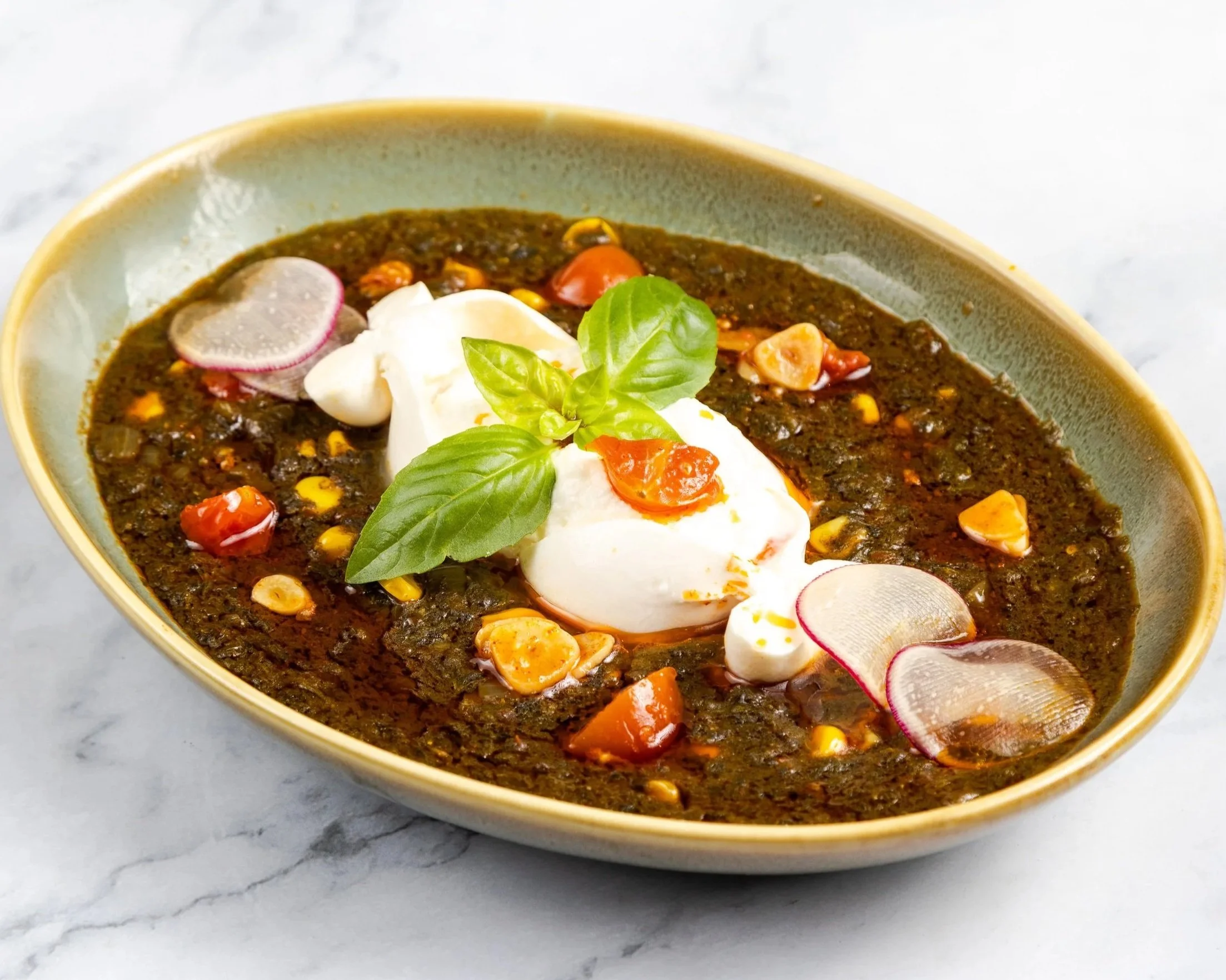 Palak preparation garnished with burrata ball, fresh basil leaves, sliced radishes, cherry tomatoes, and corn kernels, placed on a white marble surface.