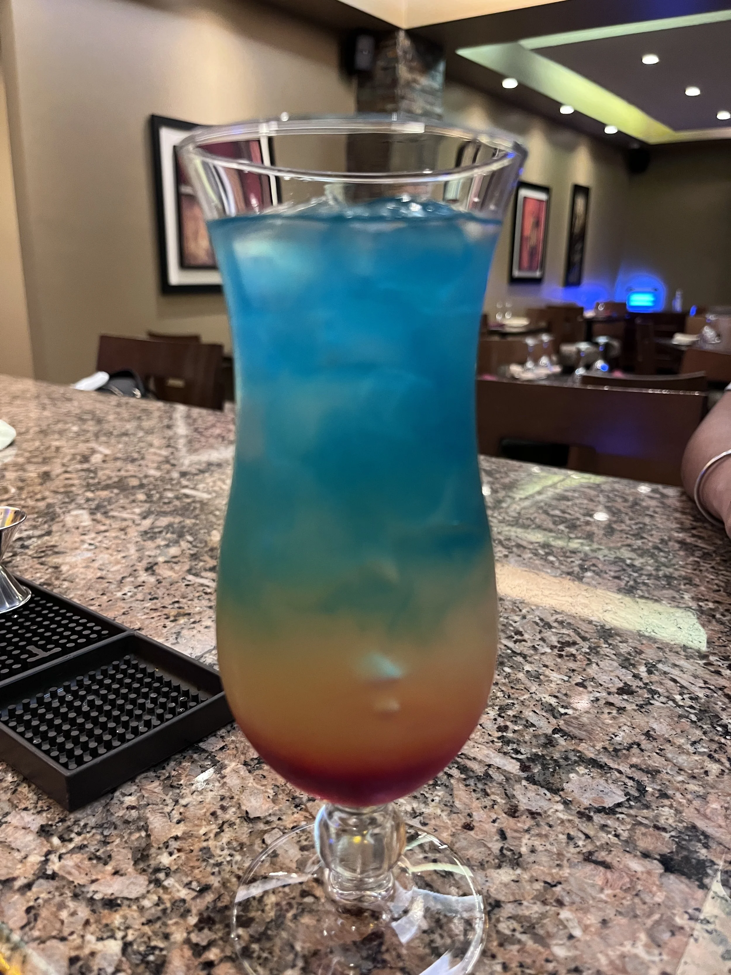 Colorful layered cocktail with blue, yellow, and red hues in a tall glass on a bar counter.