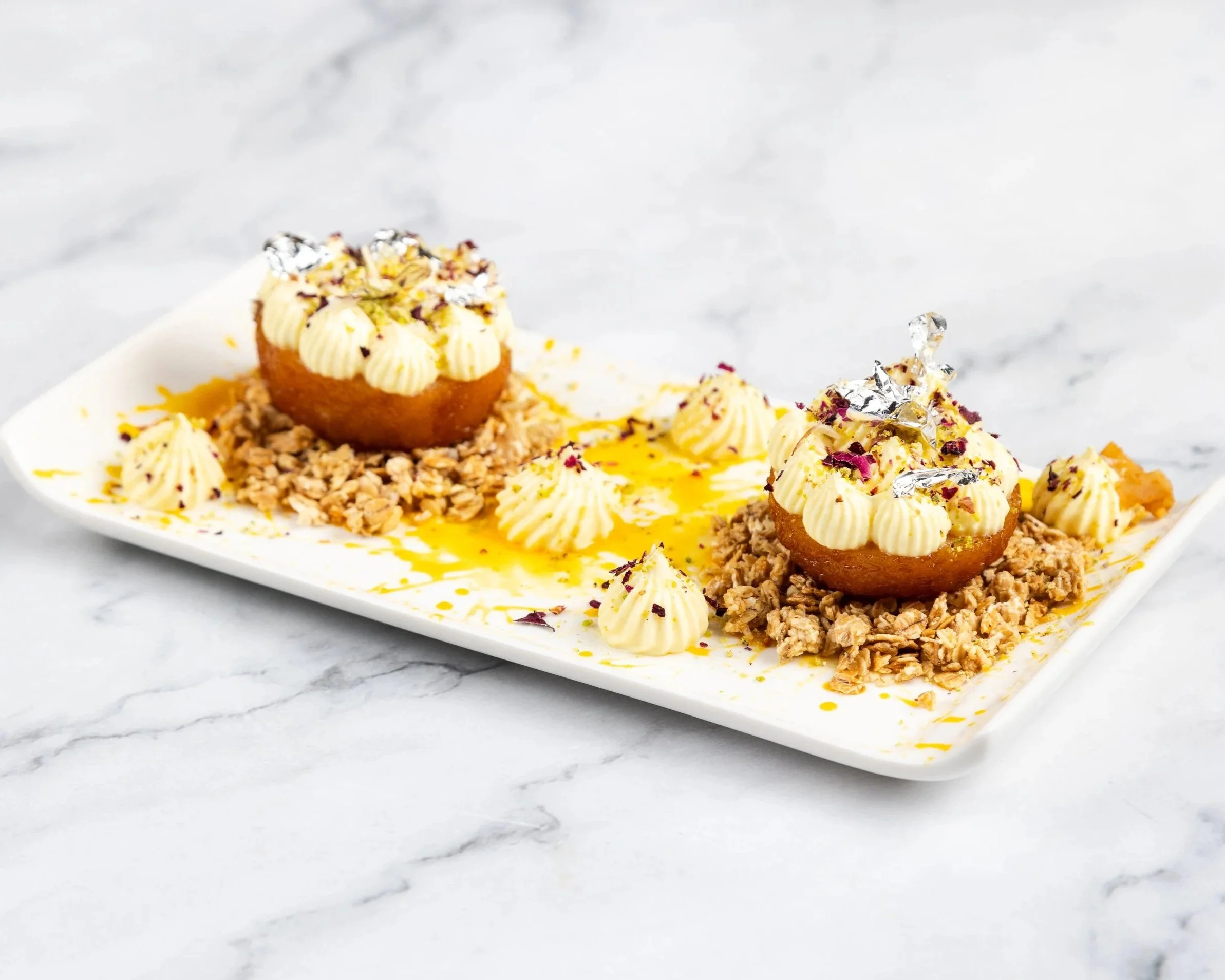 Gulab Jamun balls, topped with edible silver leaf, placed on a bed of granola on a white plate.