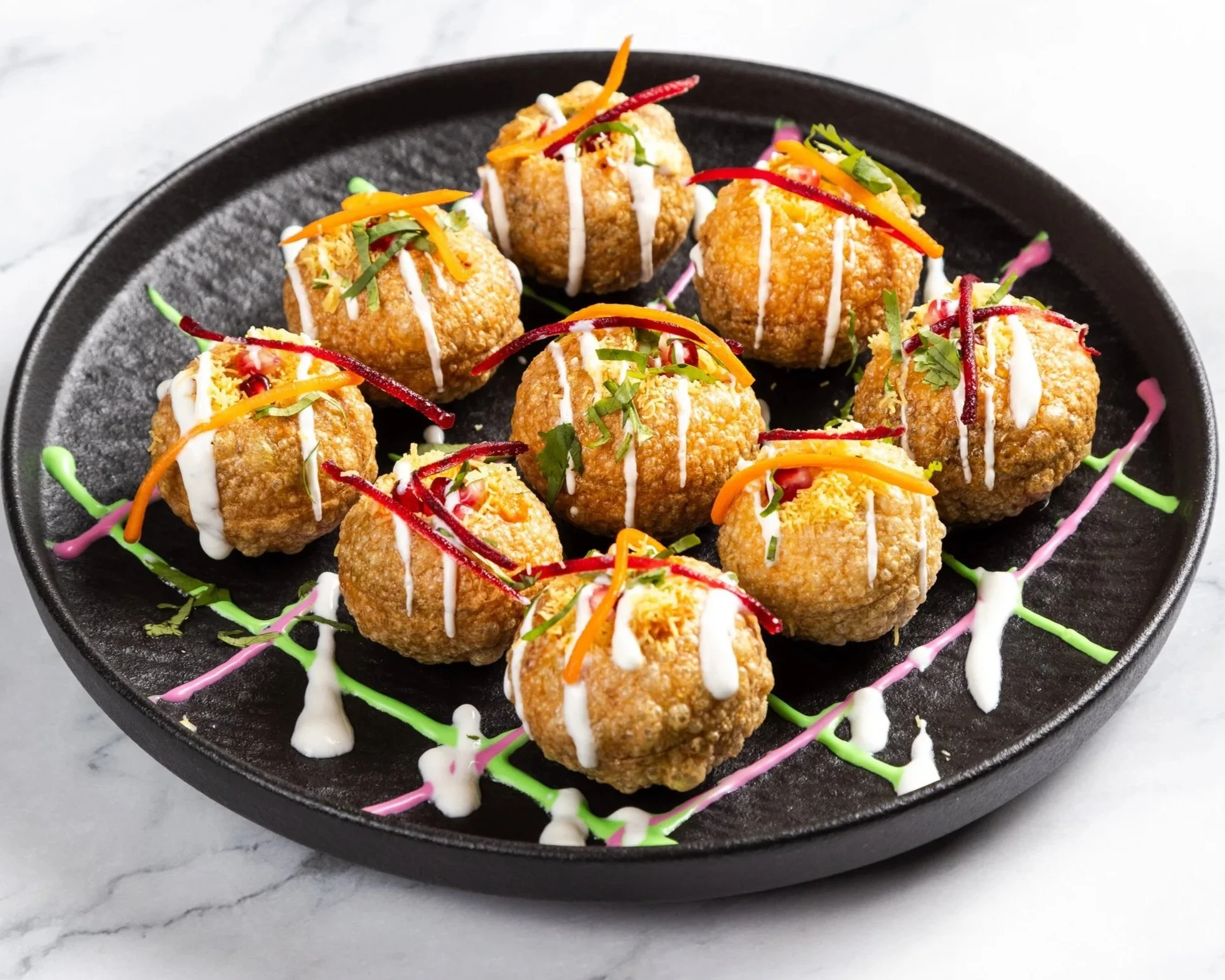 Plate of fried snack balls decorated with colorful sauce, shredded vegetables, and herbs on a black round plate.