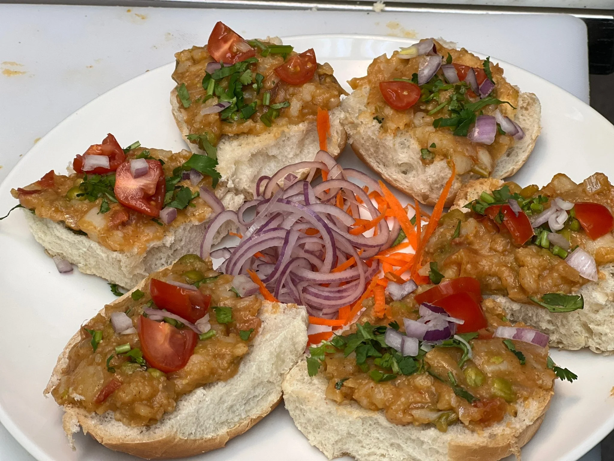 Open-faced sandwiches topped with mashed vegetables, chopped tomatoes, onions, and cilantro, with sliced red onions and shredded carrots in the center on a white plate.