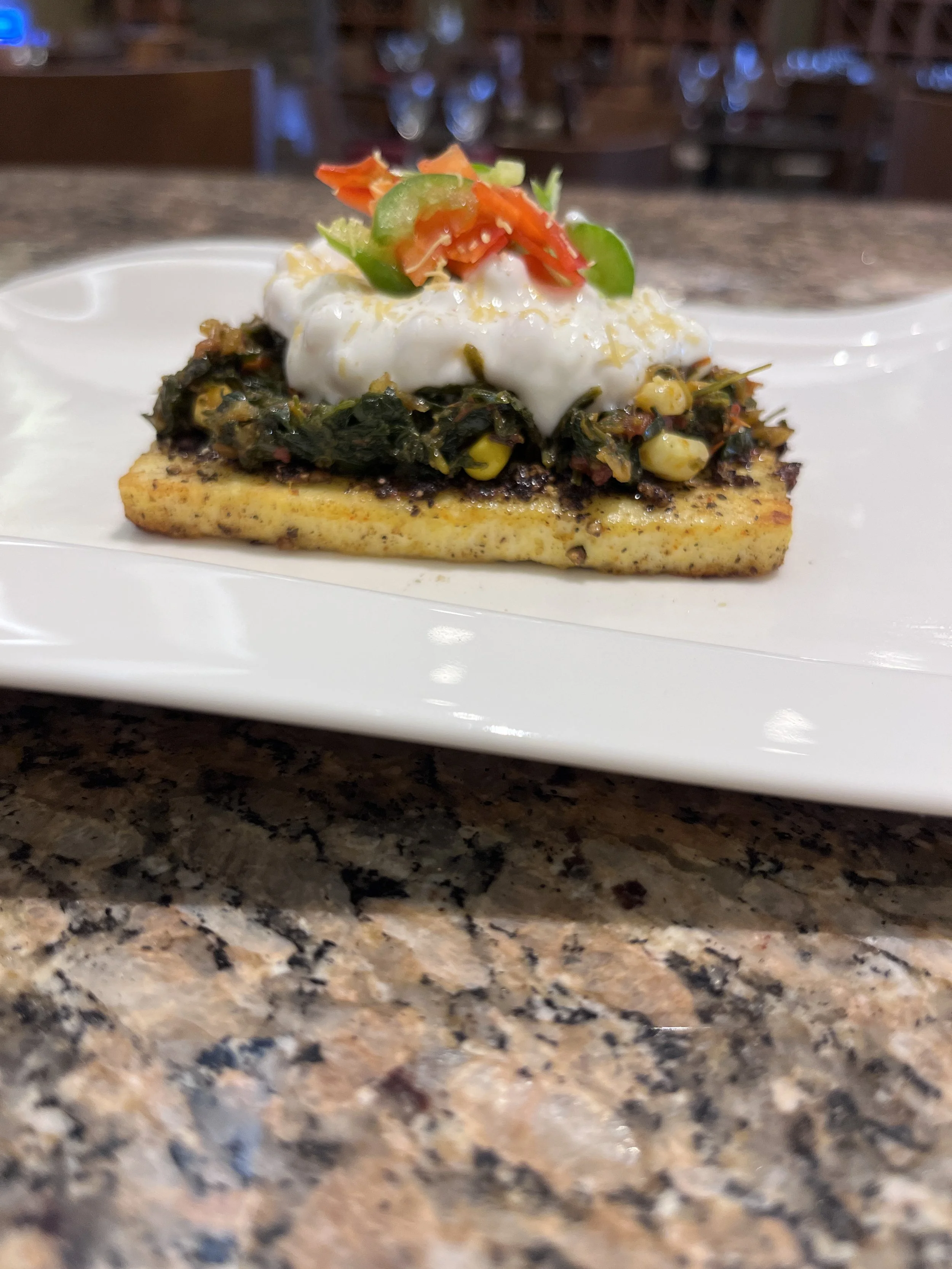 Gourmet appetizer on a white plate with spinach, corn, cream, and diced peppers on a toasted base, set on a granite countertop.