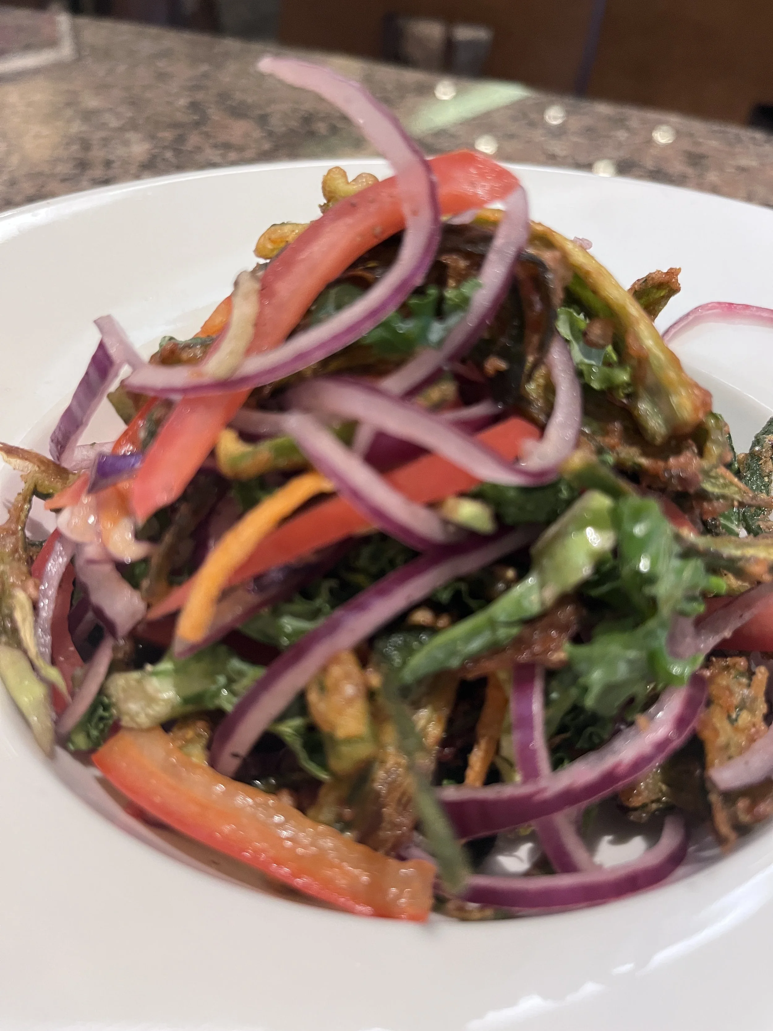 Close-up of a fresh salad with sliced red onions, red bell peppers, greens, and possibly fried elements in a white bowl on a brown countertop.