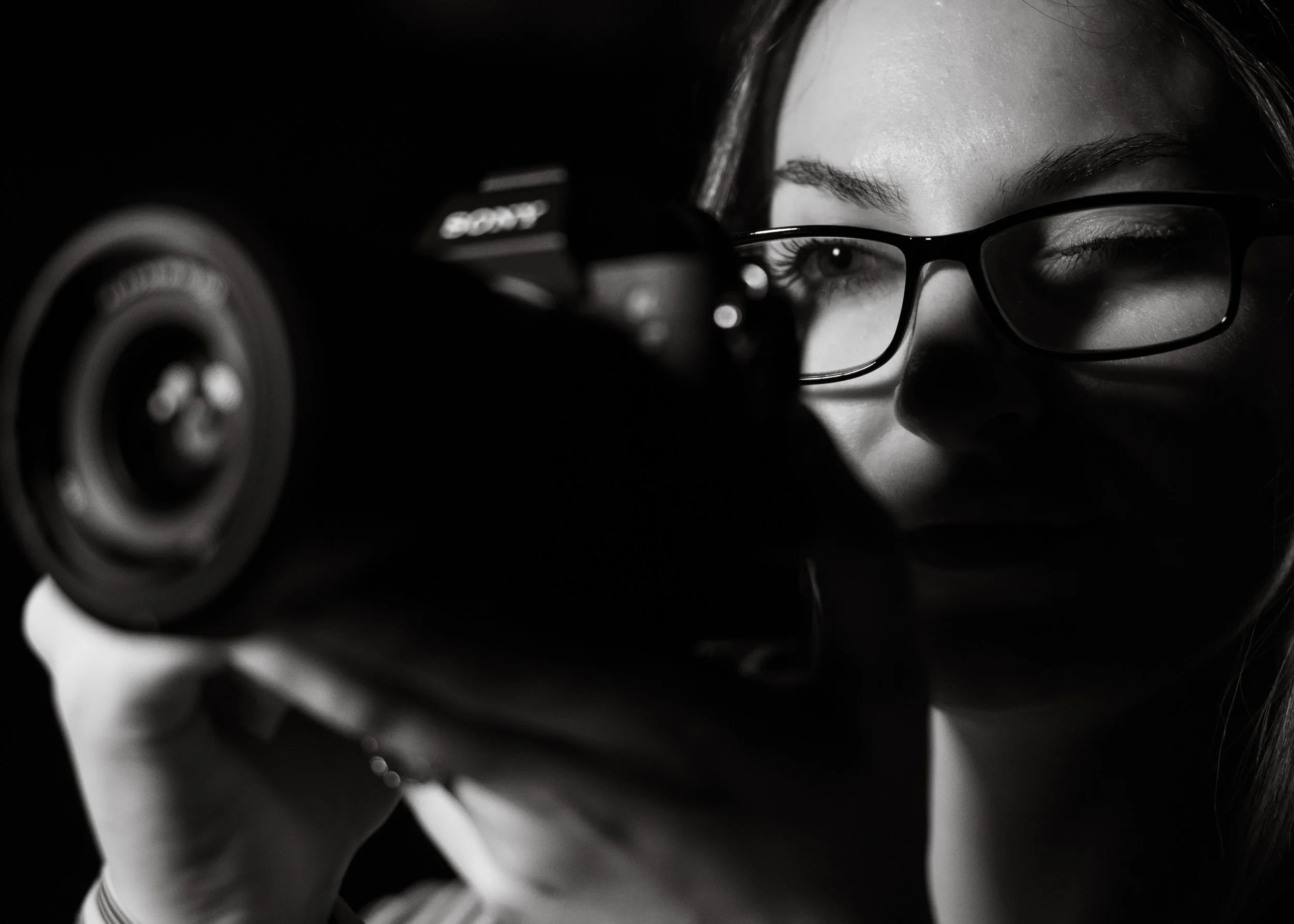 Black and white close-up of a person with glasses holding a camera, focusing on the lens.