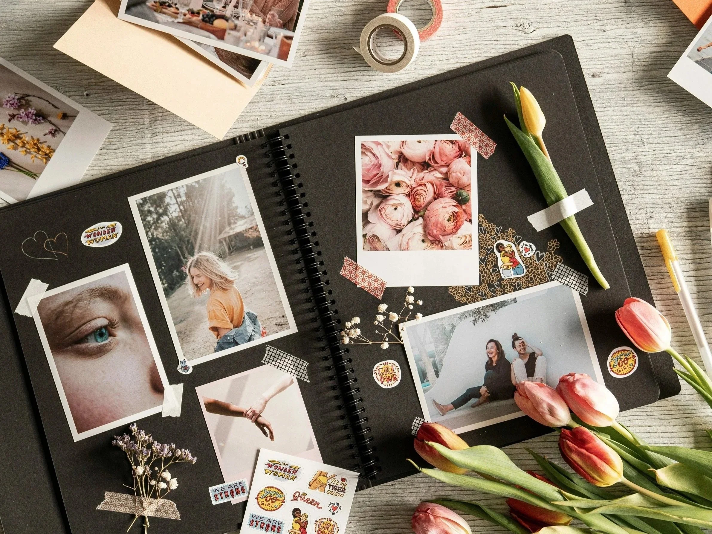 Open scrapbook on a wooden table with black pages decorated using photos, dried flowers, washi tape, and stickers. Includes tulips, pens, and colorful embellishments for DIY journaling, memory keeping, and creative scrapbooking projects.