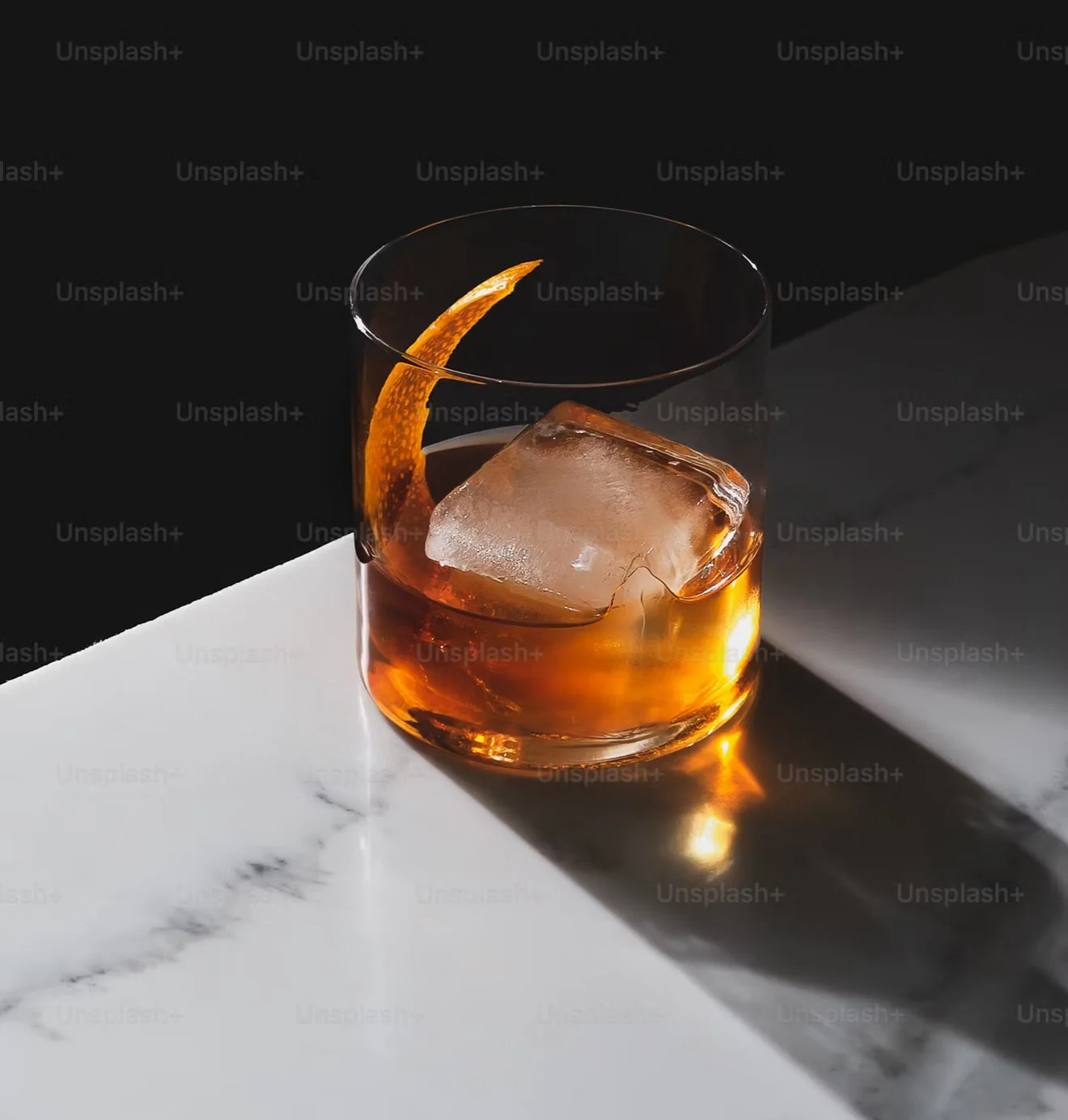 A glass of amber cocktail with a large clear ice cube and orange peel garnish sits on white marble. Soft lighting highlights the drink’s rich color and the cube’s clarity against a dark background.