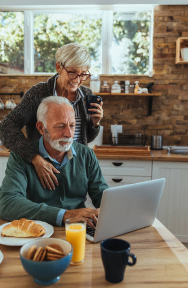 Older couple in a cozy kitchen using a laptop together. Woman smiles with coffee while man works online. Croissants and cookies on the table.