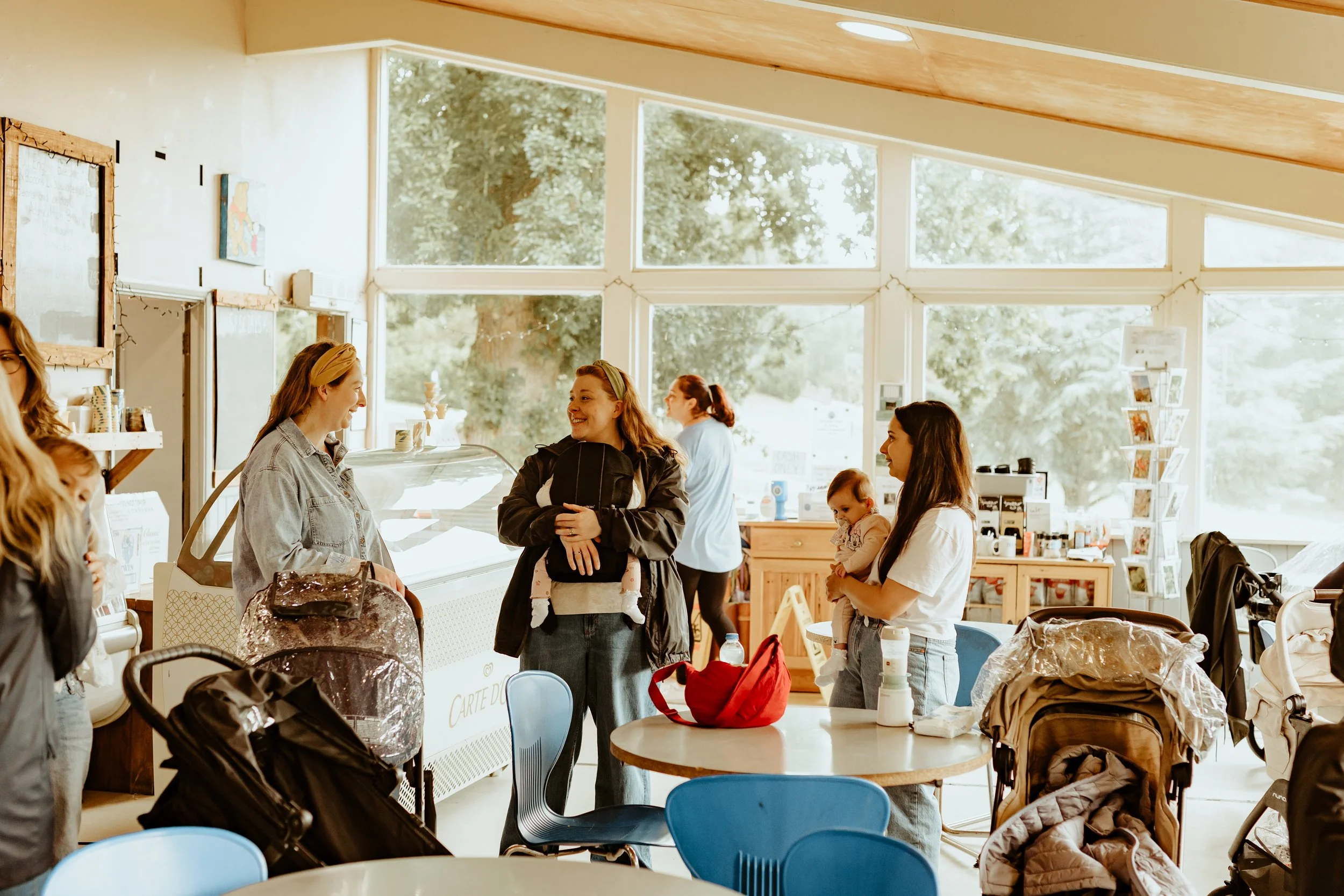 Several women and children inside a bright cafe, some with strollers, talking and smiling, with large windows revealing green trees outside.