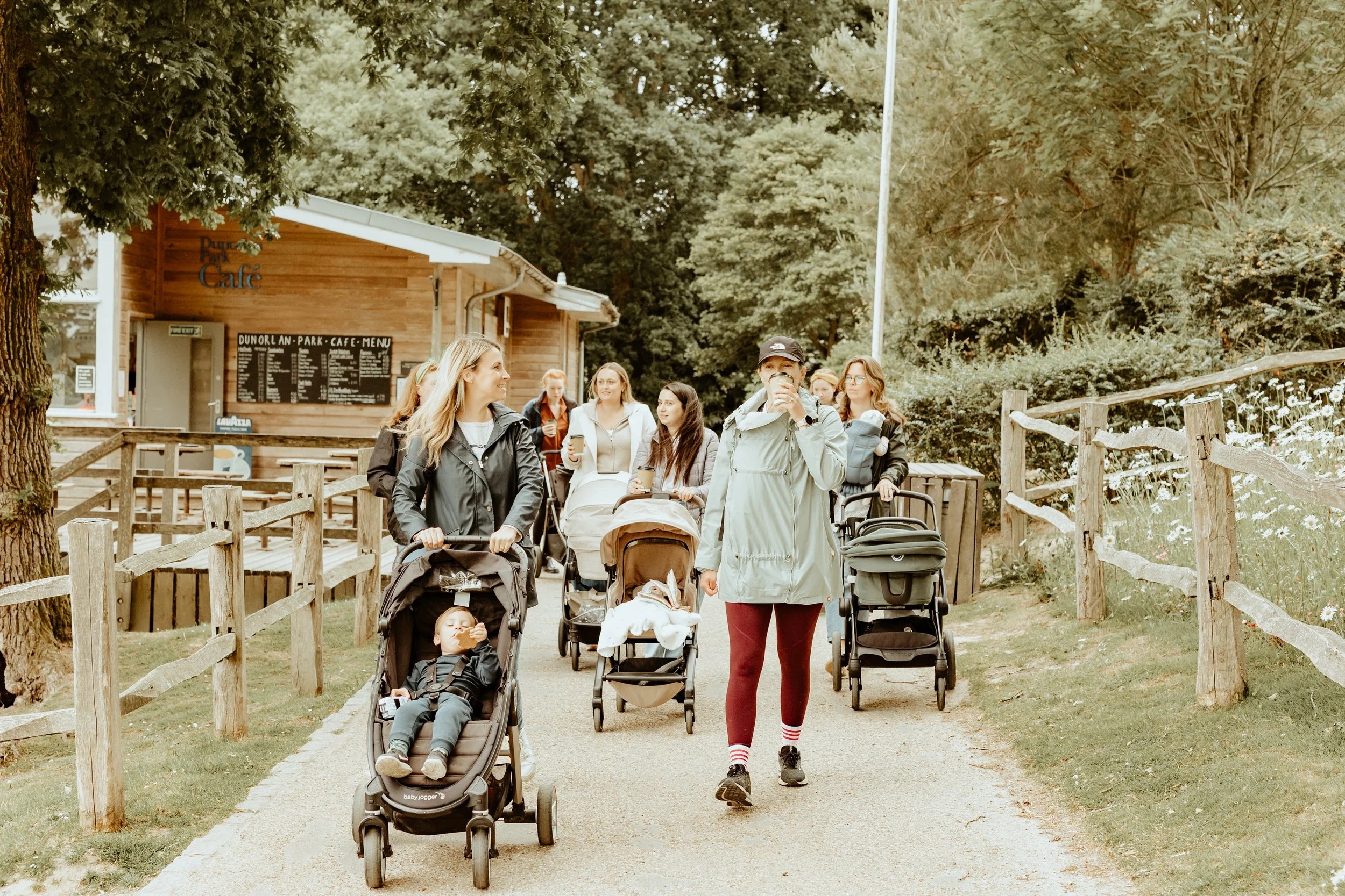 A group of women and children walking along a park path with strollers and drinks near a cafe, surrounded by trees and greenery.