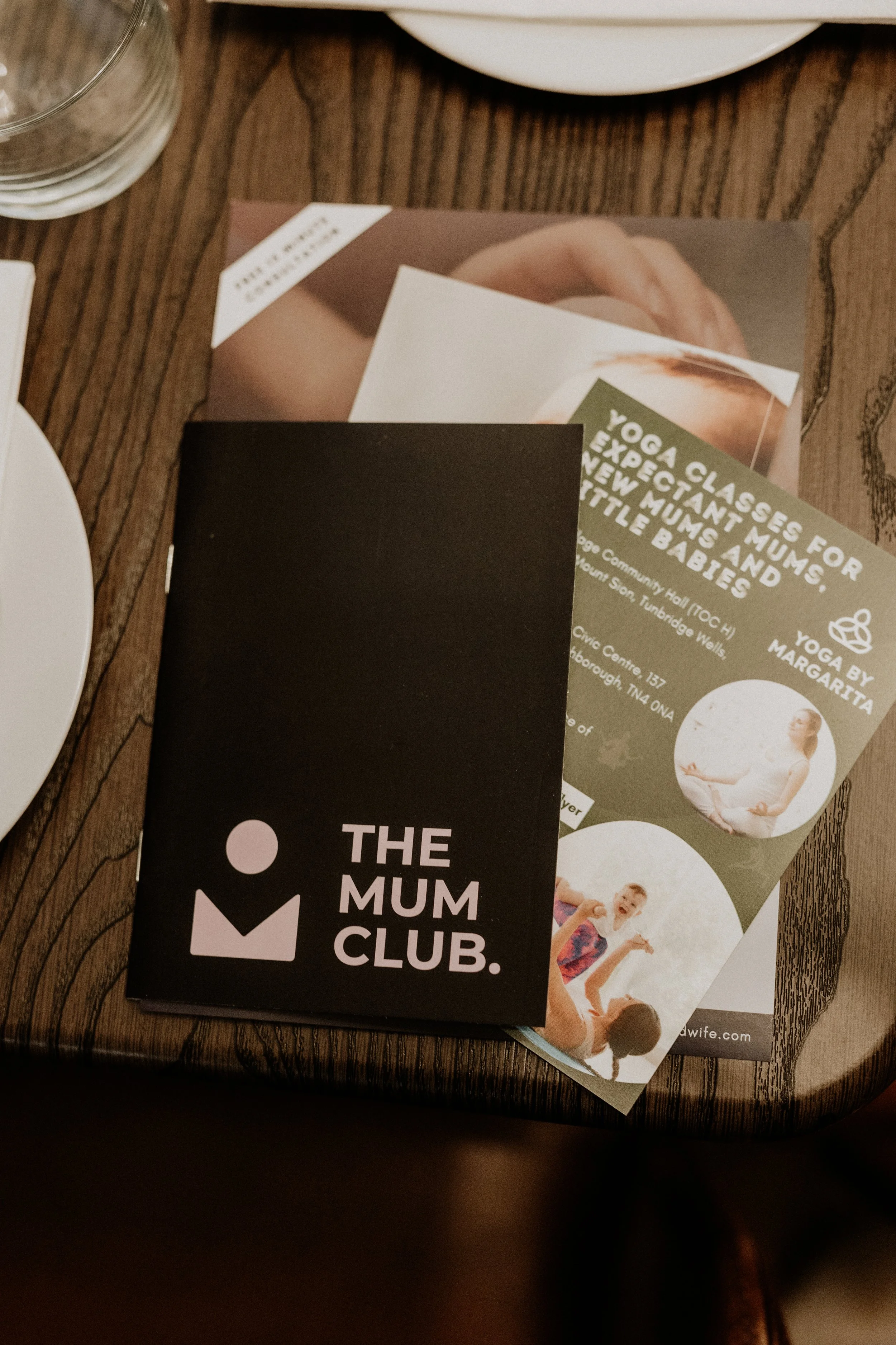 Black business card with white text reading "The Mum Club" on a wooden table, partly covering a flyer promoting yoga classes for new mums and babies.