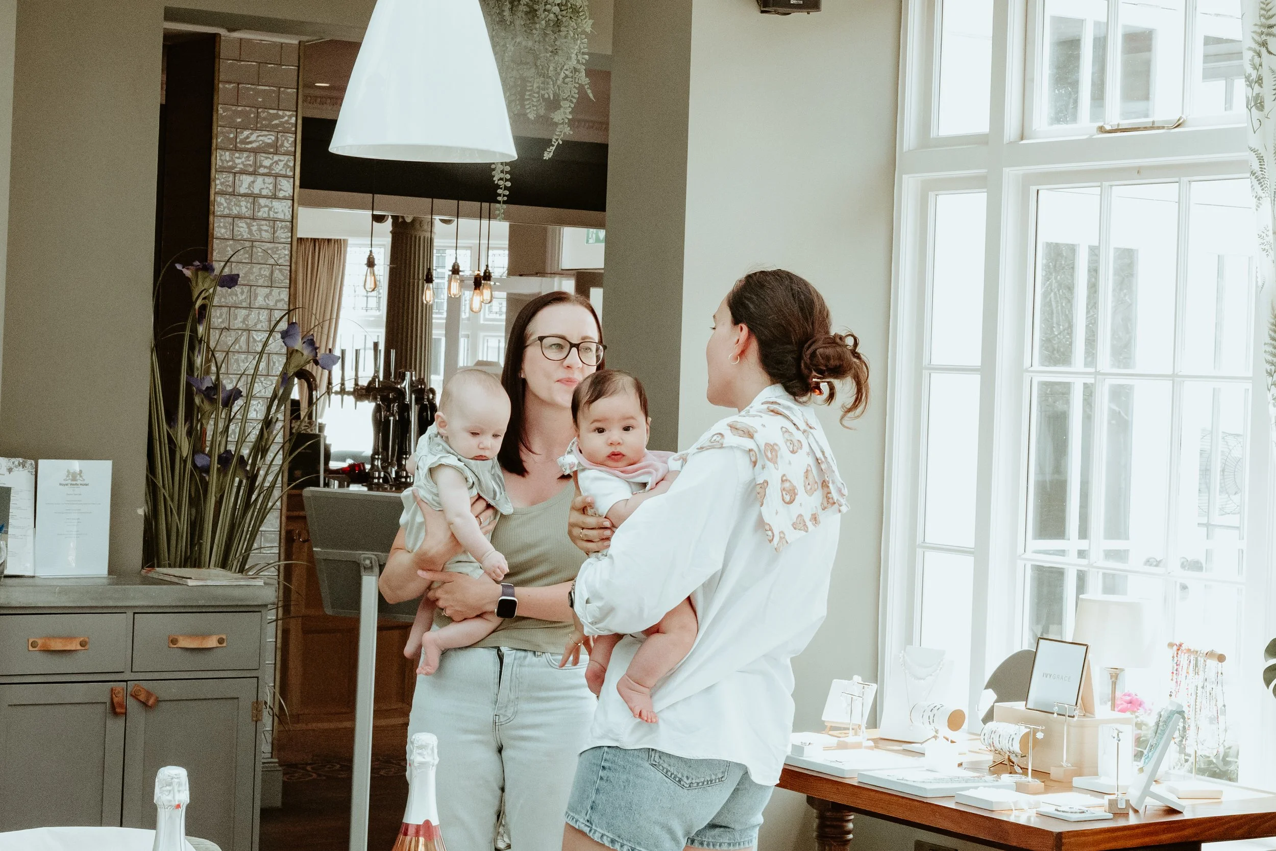 Two women holding babies in a bright, modern room with large windows and table with jewelry displays.