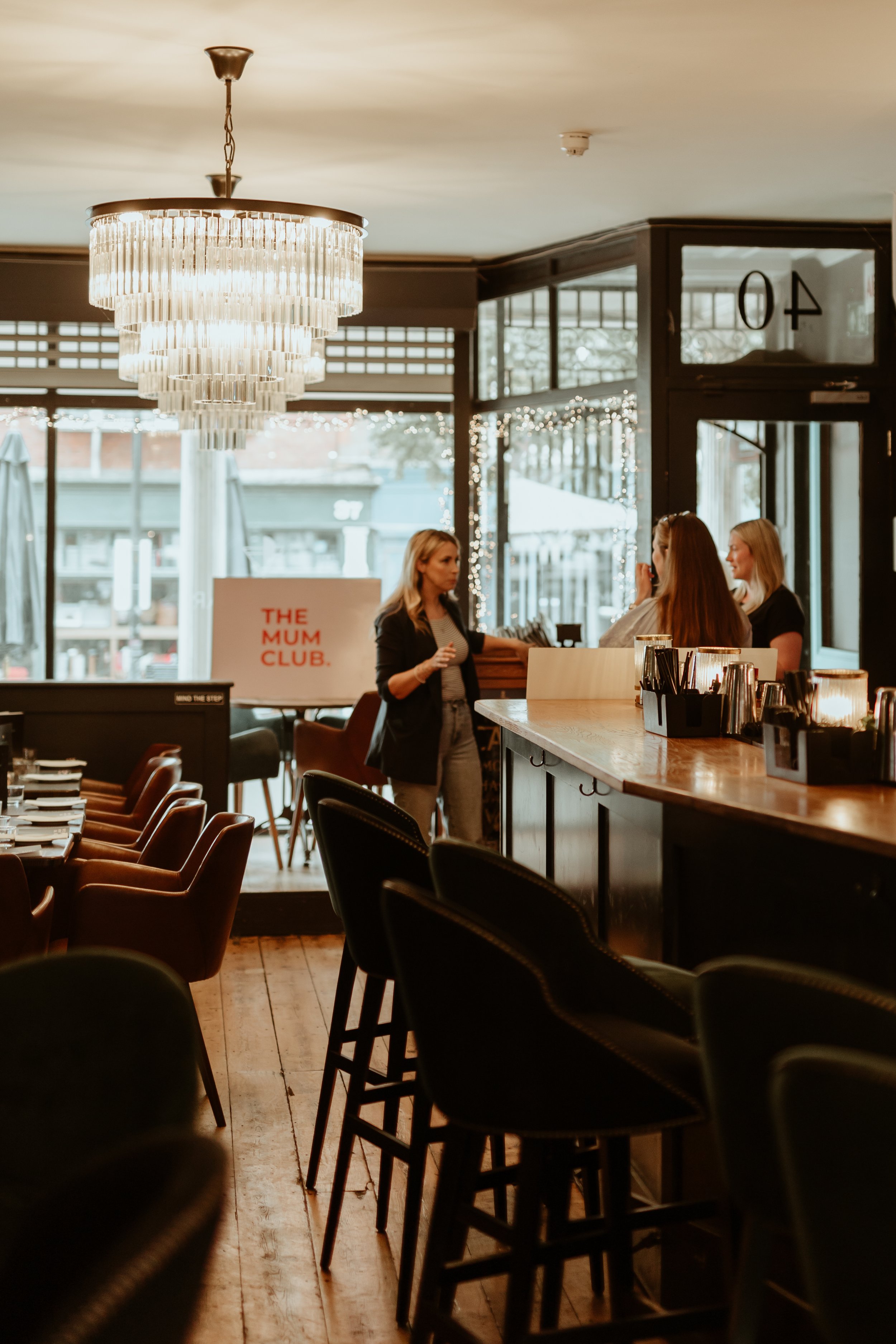 Interior of a restaurant or cafe with a chandelier, bar seating, and customers talking at the bar. There is a sign that reads 'The Mum Club' near the window.