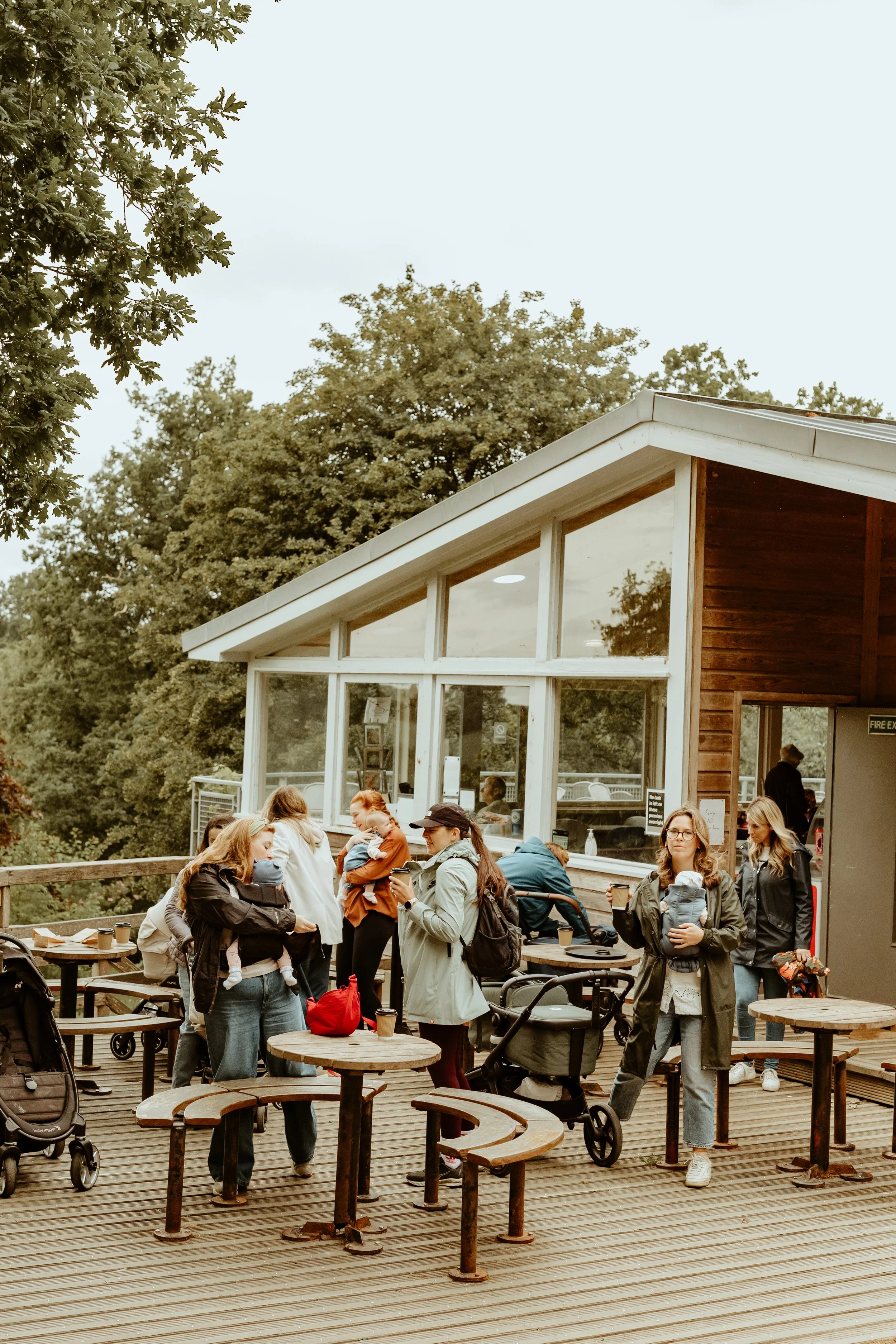 People waiting outside a modern café with large glass windows, wooden exterior, and outdoor seating on a deck, surrounded by trees.