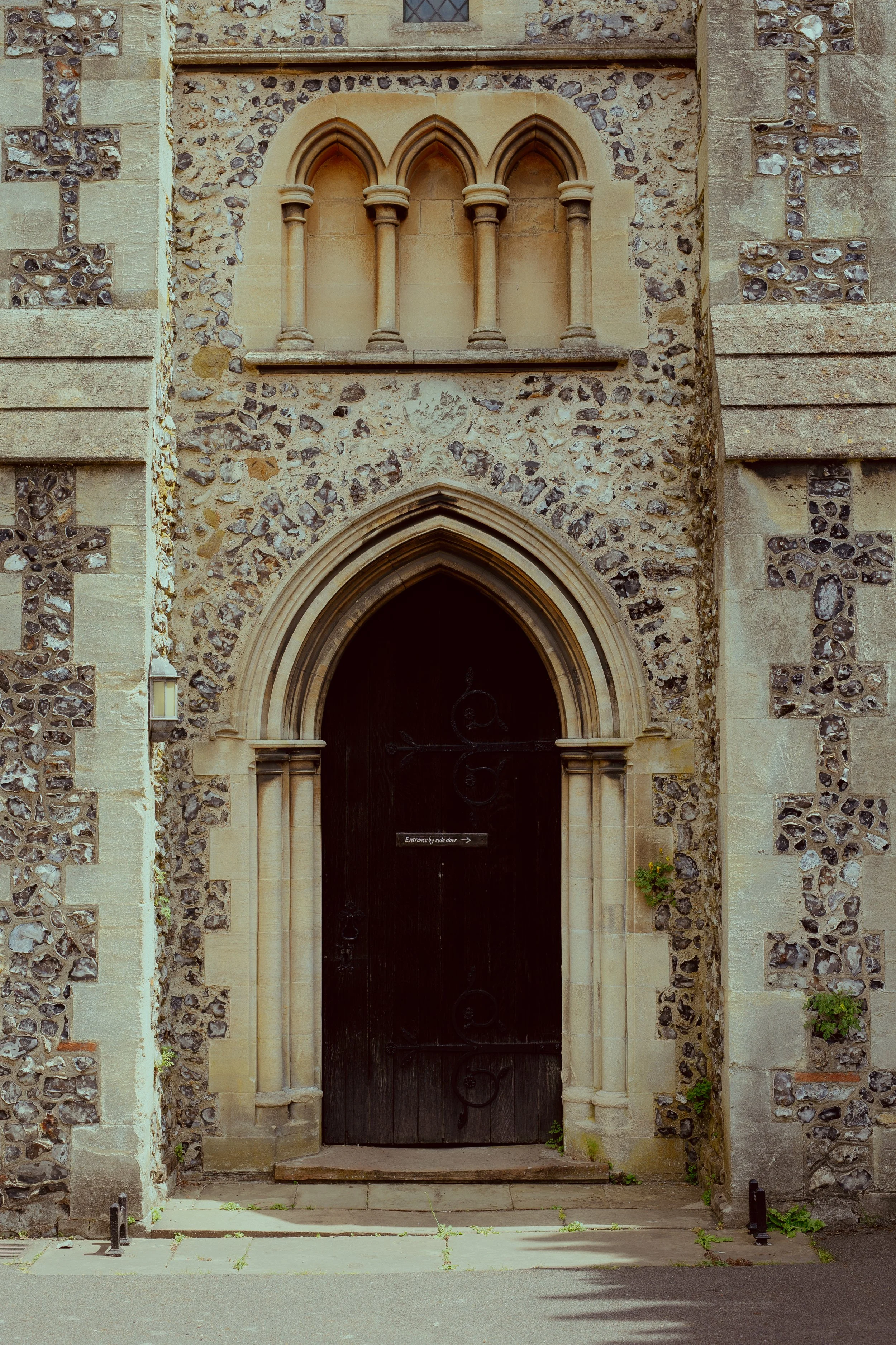 Stone church entrance with arched black wooden door and Gothic-style archway, flanked by stone columns and decorative stonework, with a small window above.