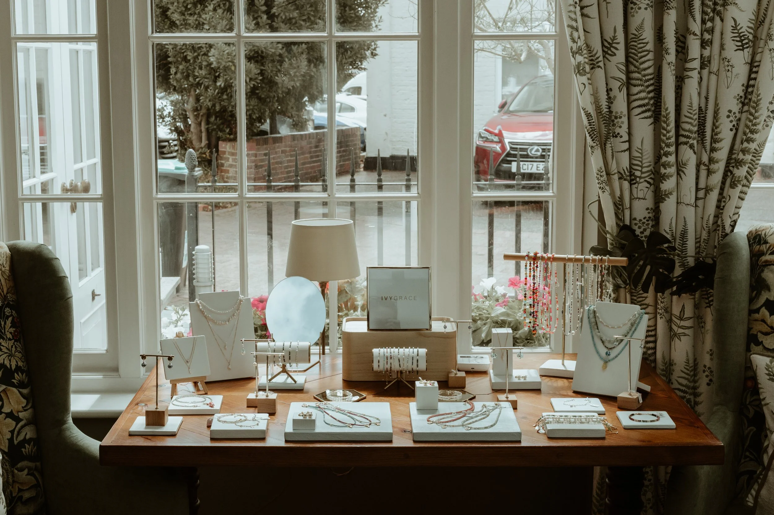 Jewelry display table with necklaces, bracelets, and earrings in front of a window with curtains, a lamp, and outdoor cars.