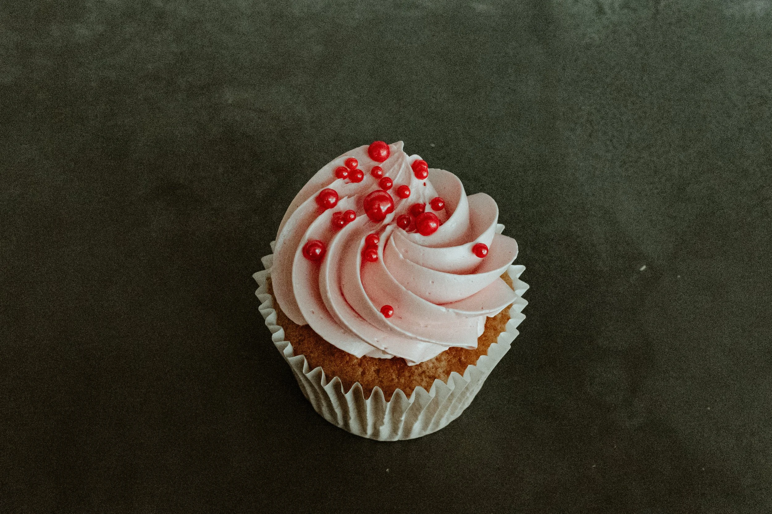A cupcake with pink frosting decorated with red sugar pearls on a dark surface.