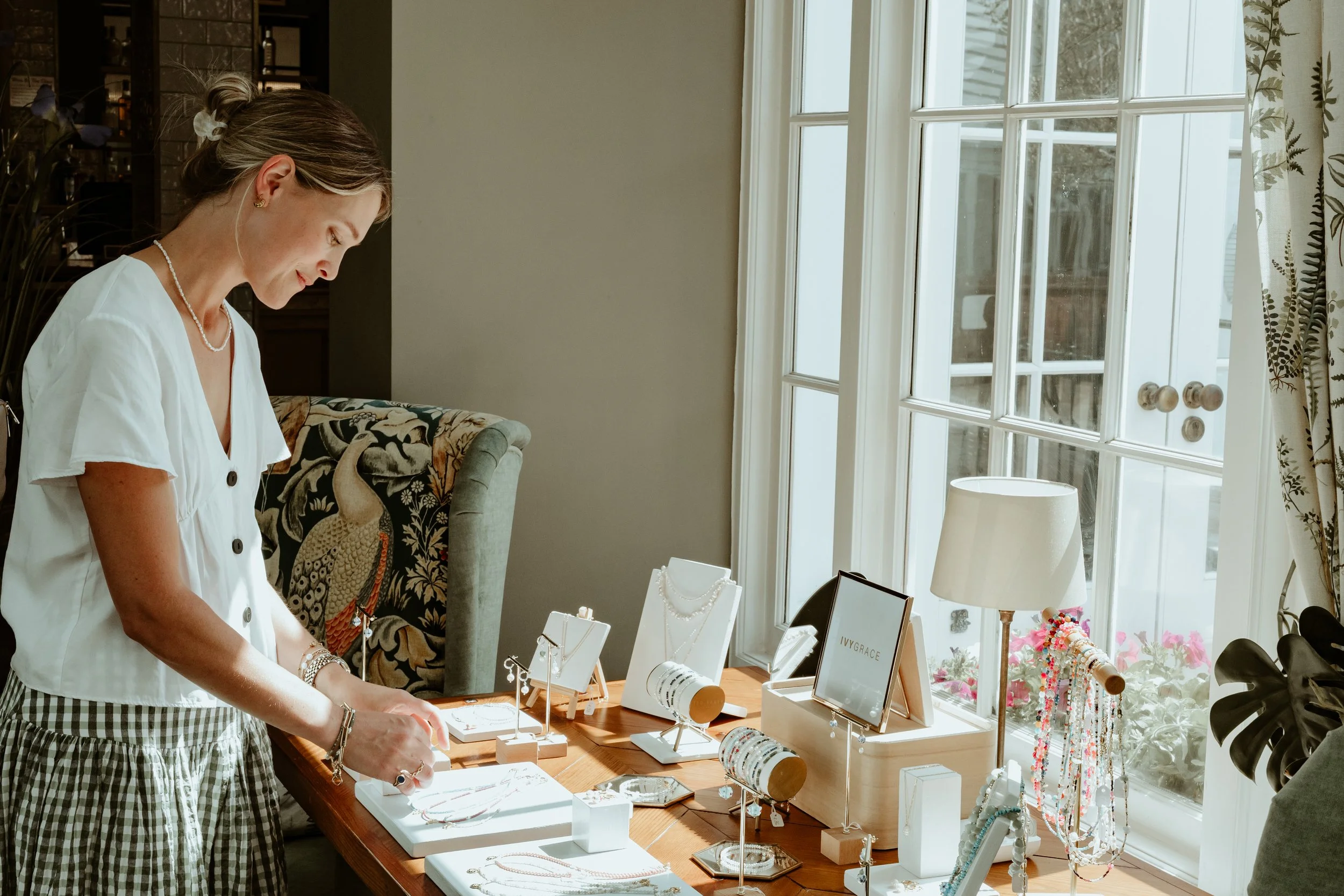 Woman browsing jewelry on a display table in a well-lit shop with large window.