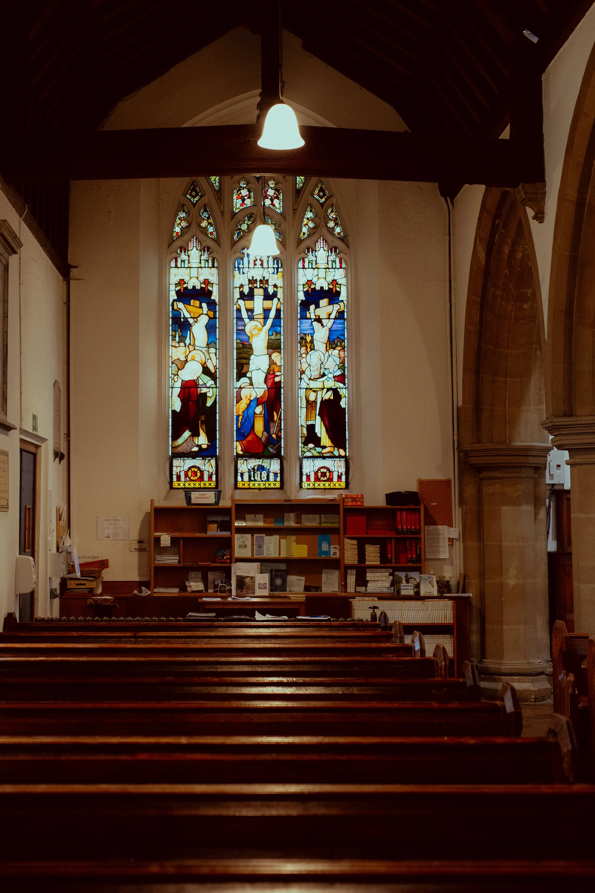 Interior of a church with wooden pews, a stained glass window depicting the crucifixion of Jesus Christ, and a hanging light fixture.