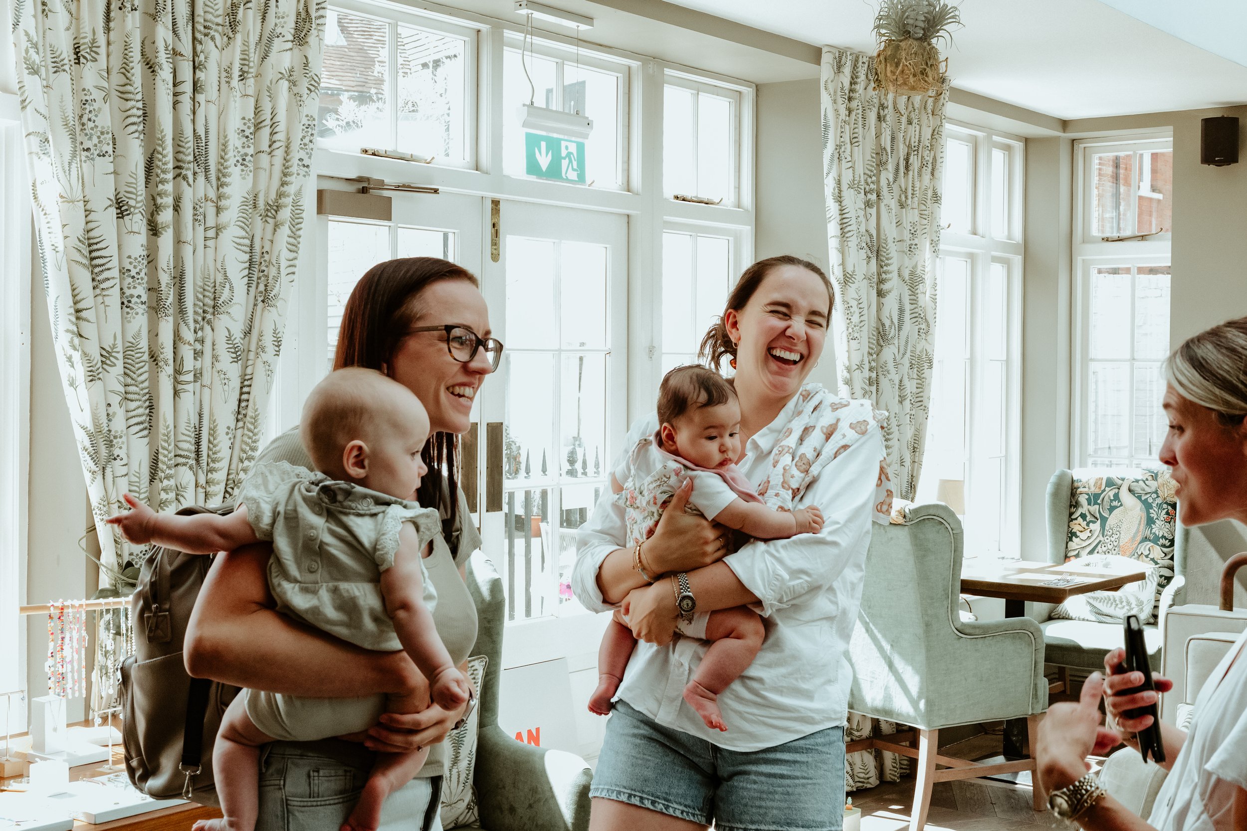 Three women holding babies and smiling in a bright, cozy room with large windows and curtains.