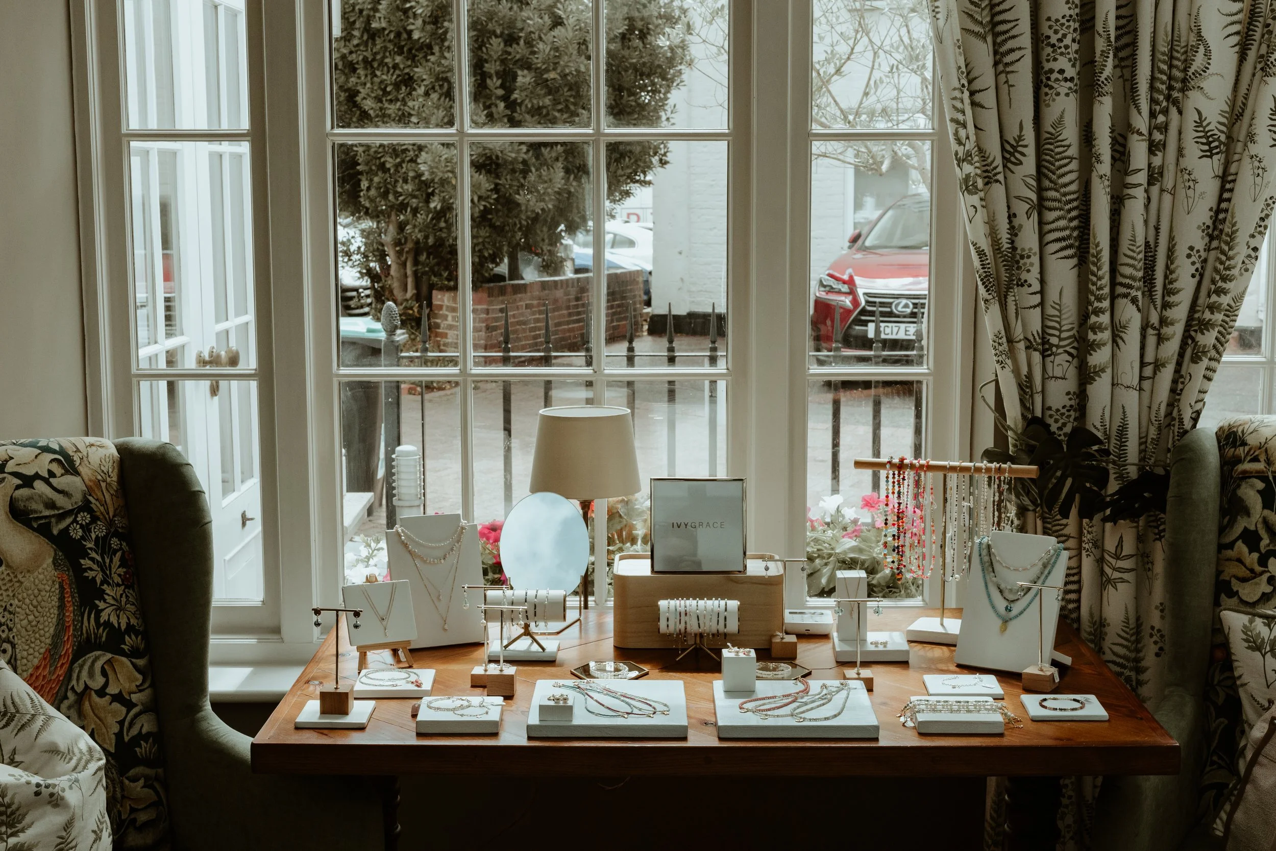 Jewelry display on a wooden table near a window with floral curtains, showcasing necklaces and bracelets in a cozy boutique setting.