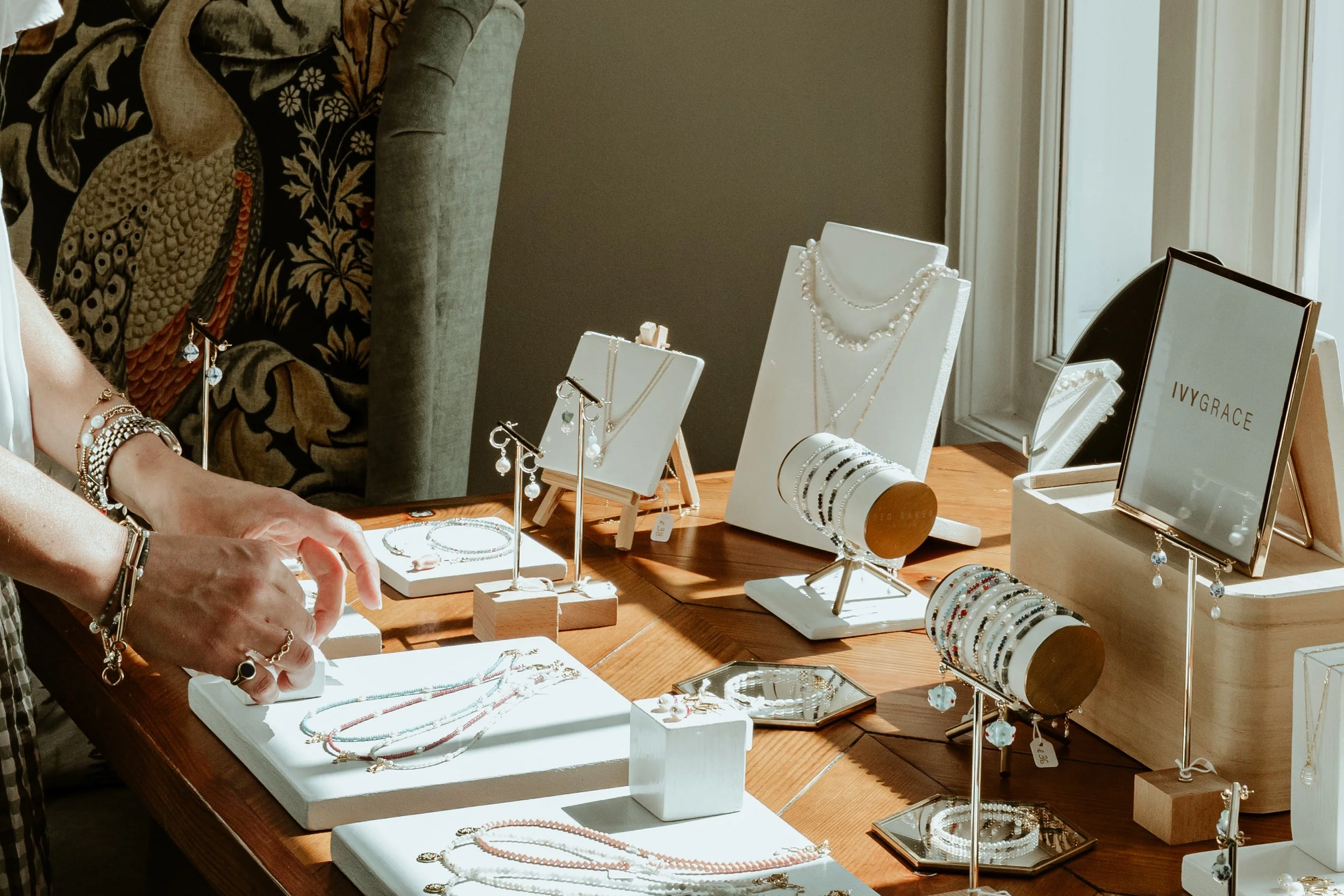 Jewelry display with necklaces, bracelets, earrings, and rings on a wooden table near a window.